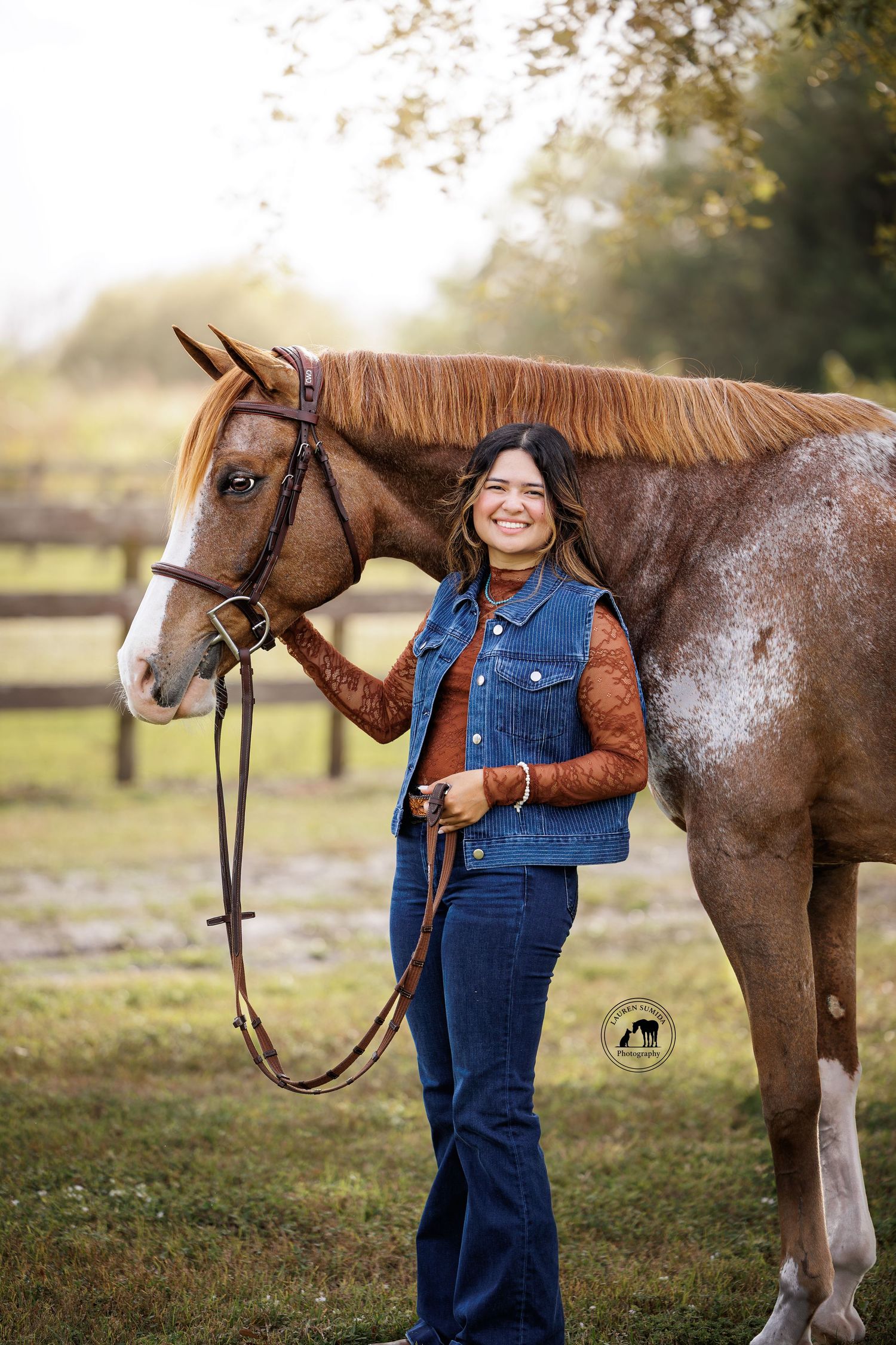 A roan horse with distinctive markings stands in a grassy field with someone in denim overalls.