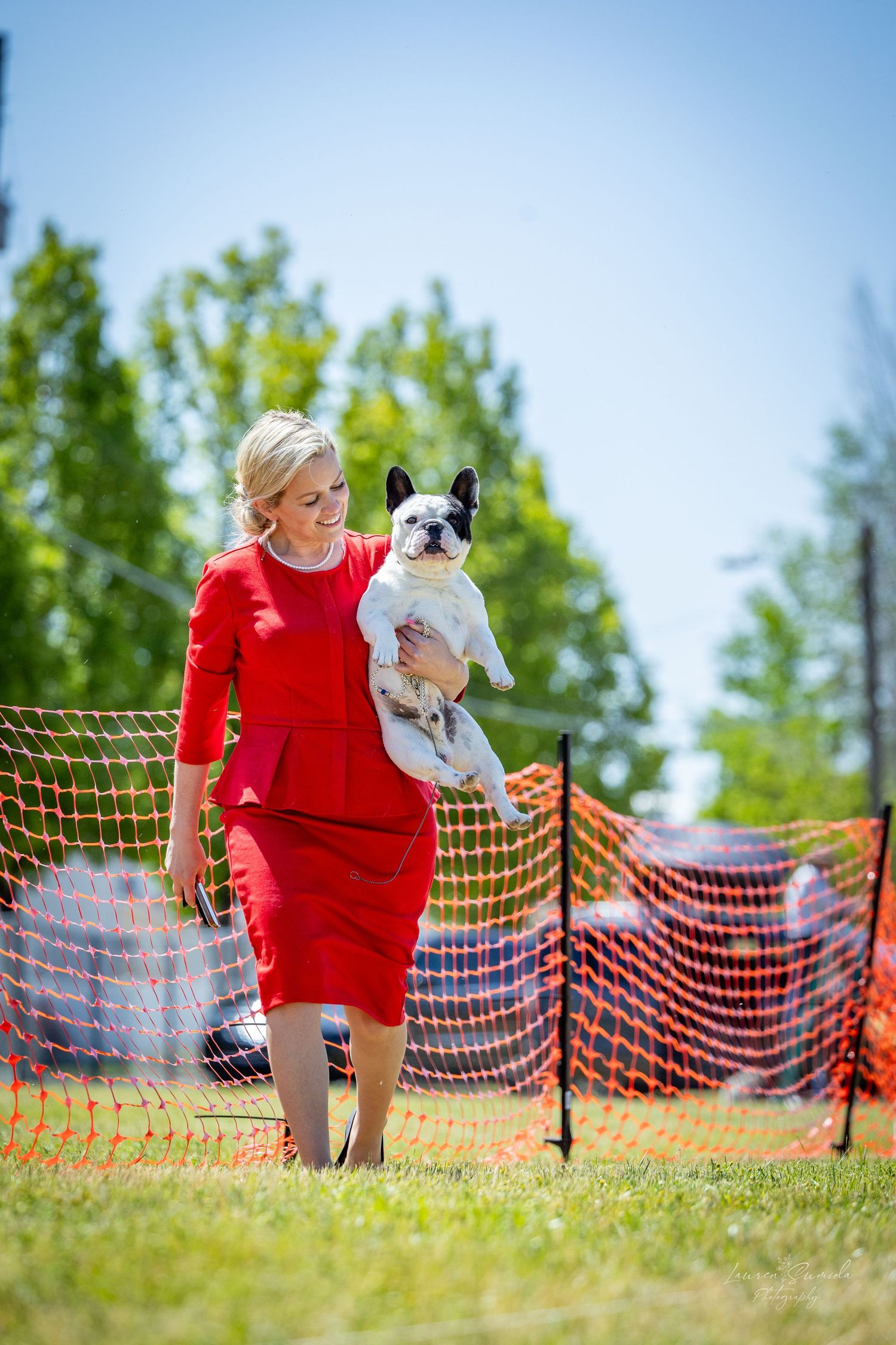 A person in red clothing plays with a small white dog on a grassy field with orange safety netting in the background.
