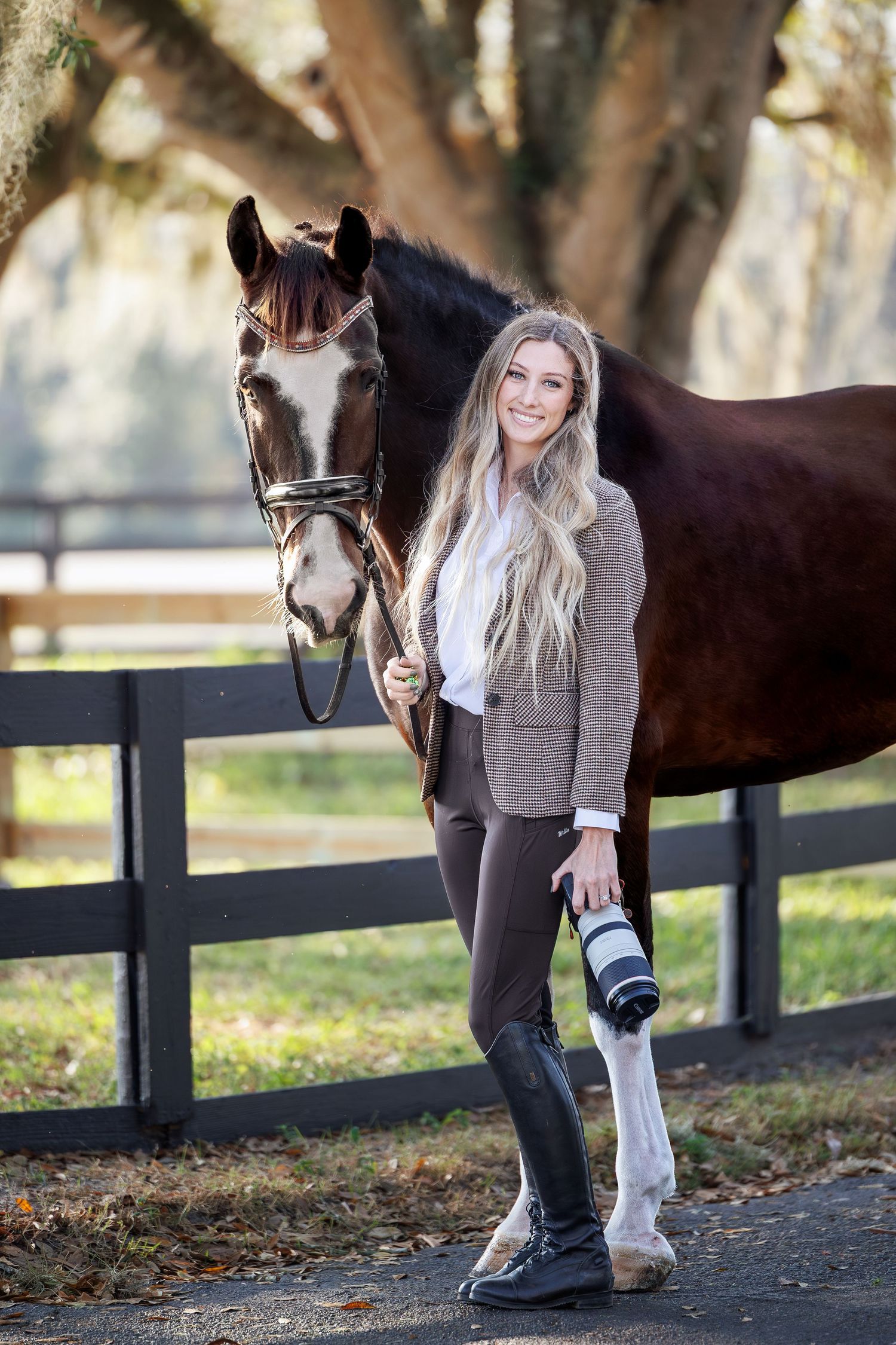 A brown horse and person stand together near a wooden fence in an autumn setting with trees in the background.
