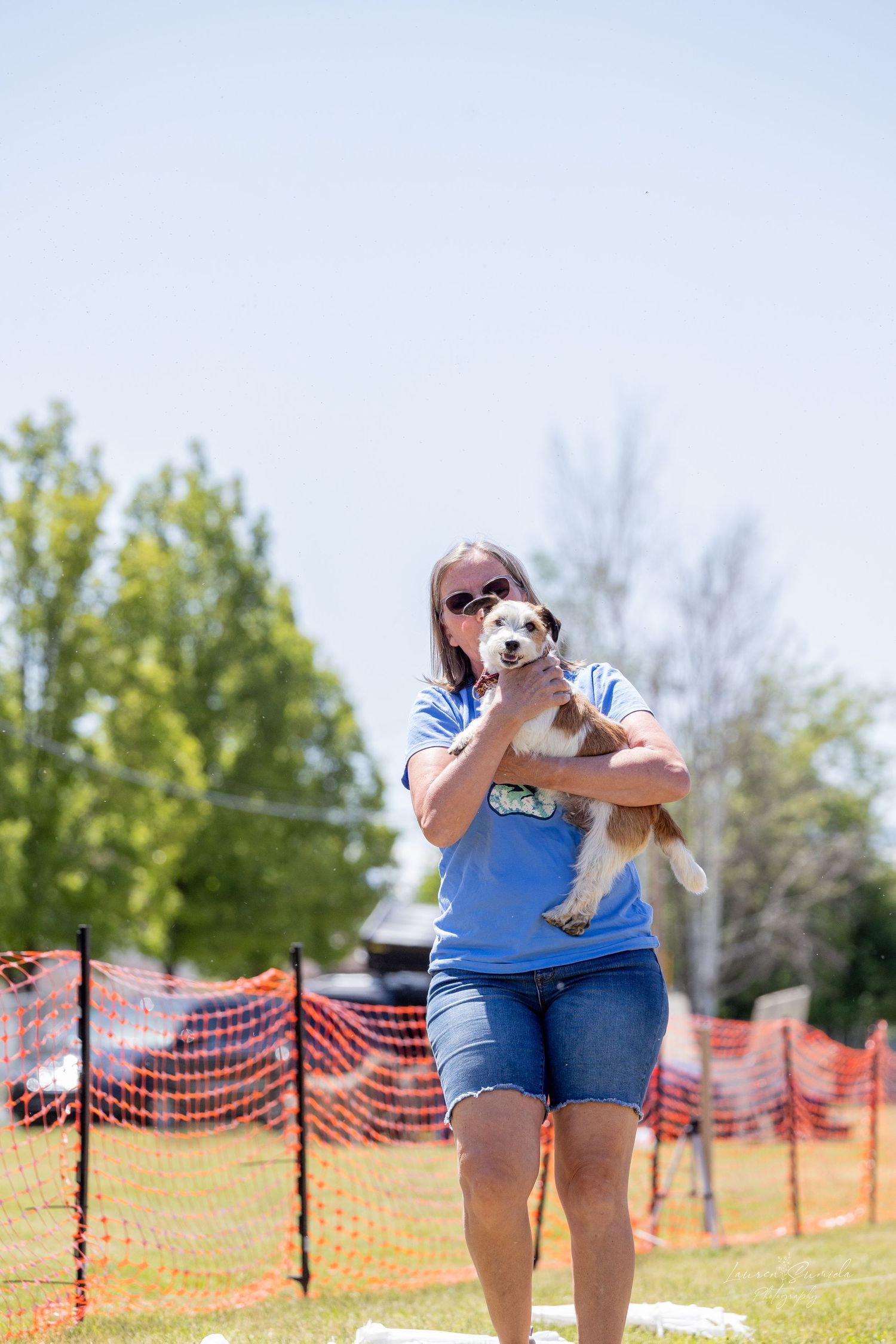 Someone in blue denim holds a small dog near orange safety fencing on a grassy field.