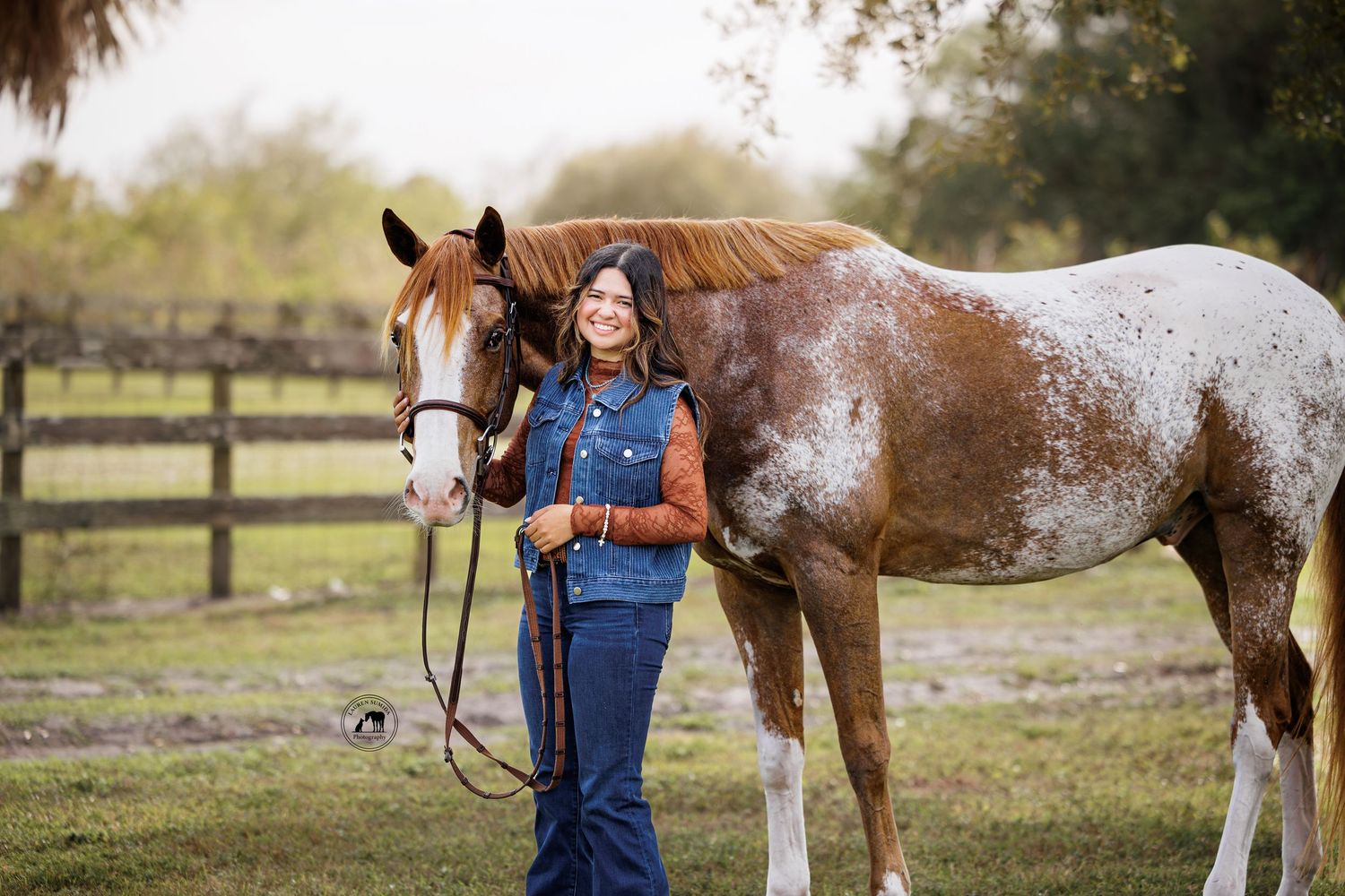 A brown and white spotted horse stands in a grassy field with someone in denim overalls and brown jacket.