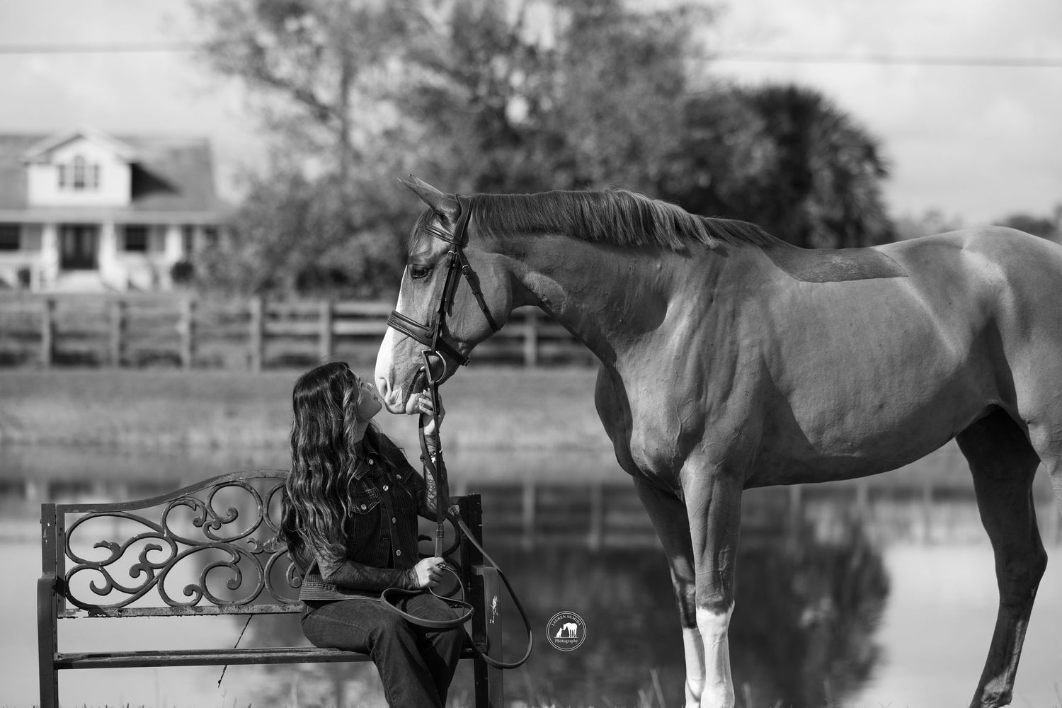 Black and white photograph of horses near water with rustic fence in background.