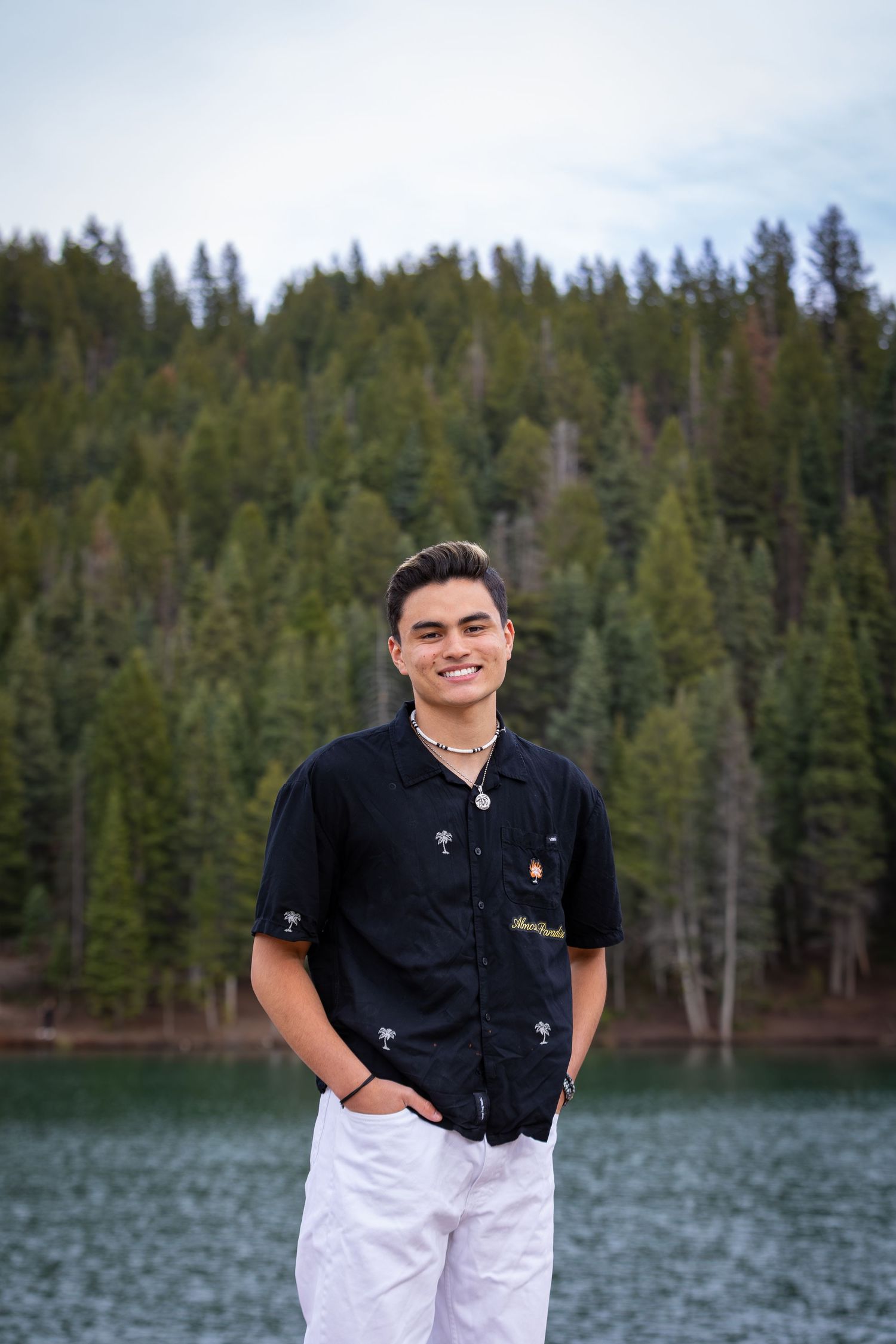Outdoor portrait session by a lake with evergreen forest backdrop.