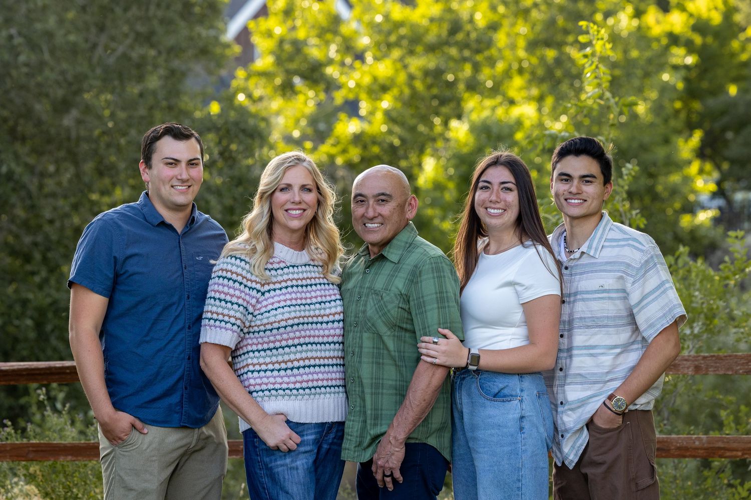 Multi-generational family smiles together on outdoor deck with sunny foliage behind them.