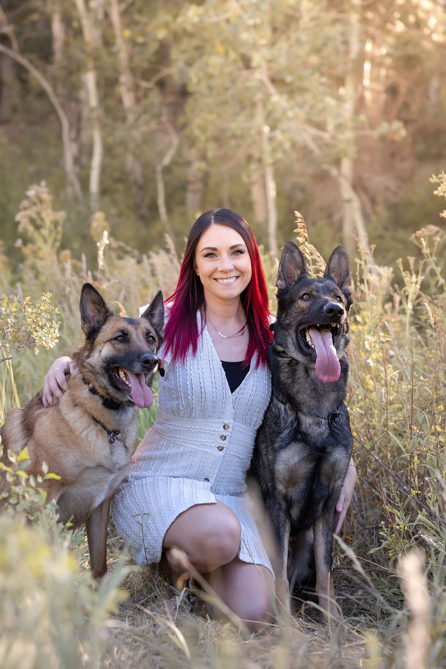 Woman with colorful hair poses with two German Shepherd dogs in tall grass at sunset.