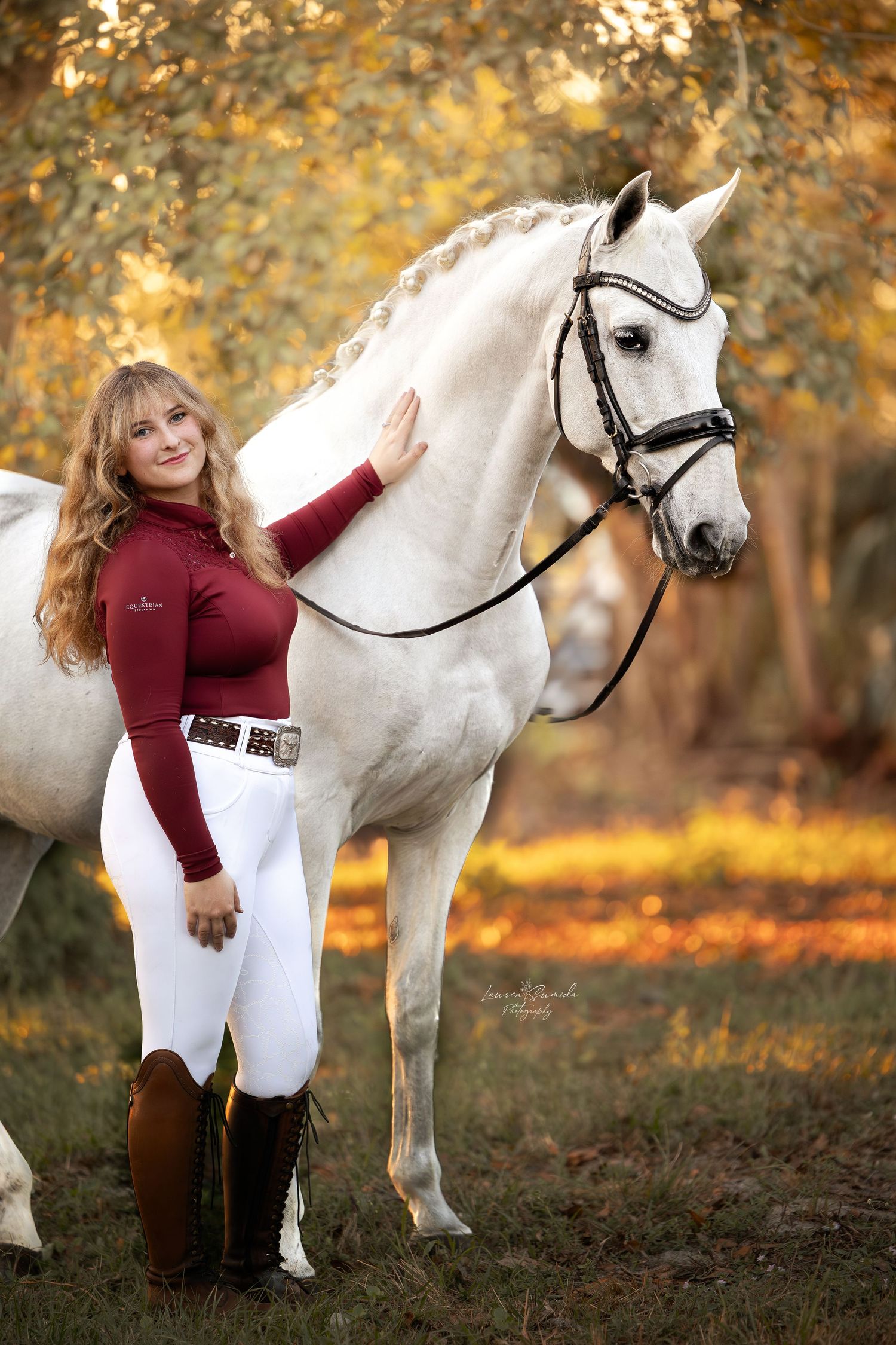 White horse with black bridle stands in autumn forest setting.