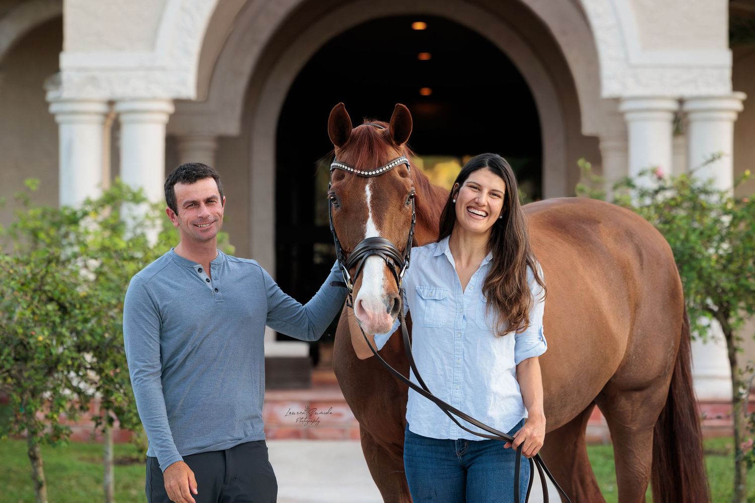 Horse stands between two people in casual attire by an arched stone walkway.