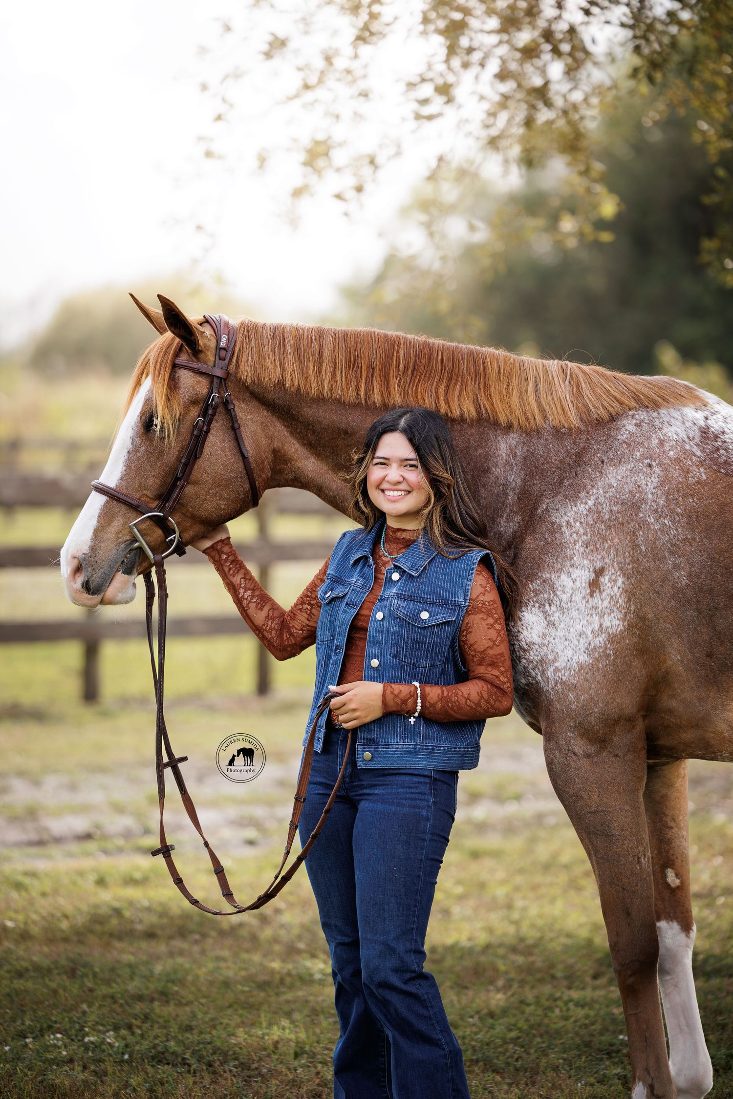 Brown and white spotted horse stands beside someone in denim overalls in sunlit field.
