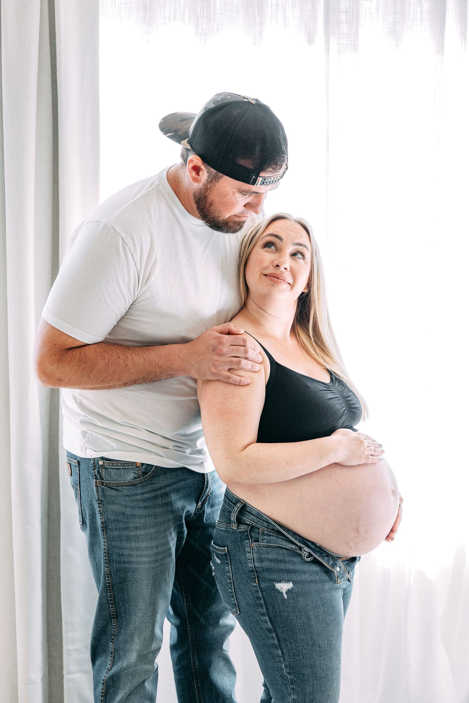 A couple embraces during a maternity photoshoot against white curtains.