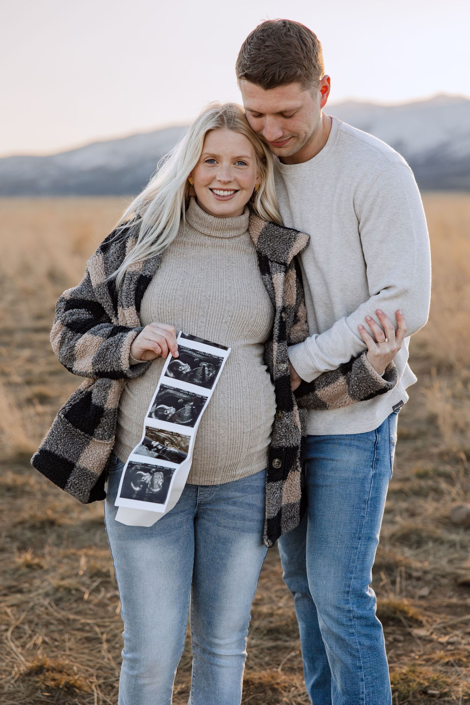 Expecting couple holds ultrasound photos in a field with mountains during sunset.