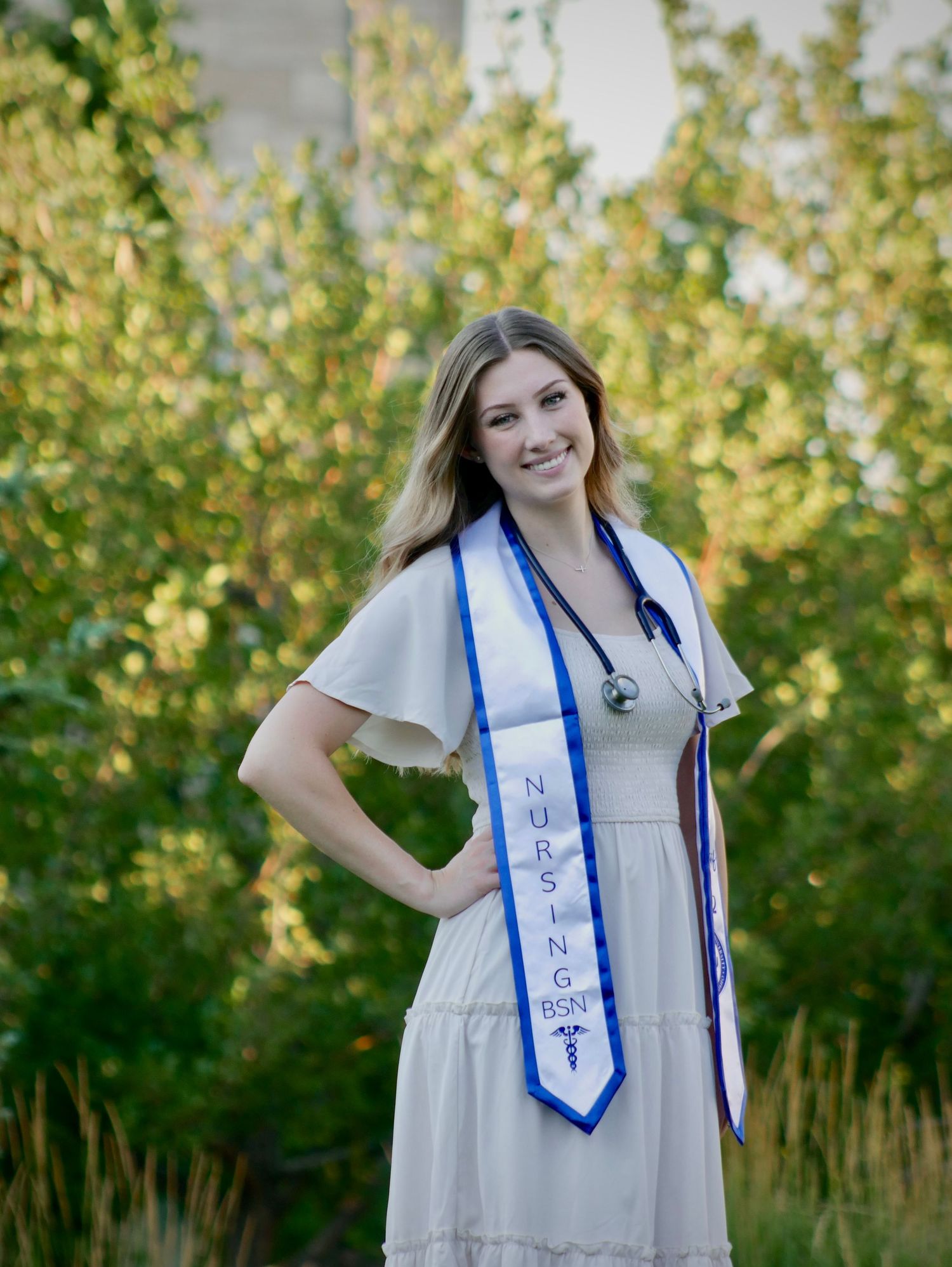 Graduate in white dress with blue graduation sash poses outdoors in front of trees.
