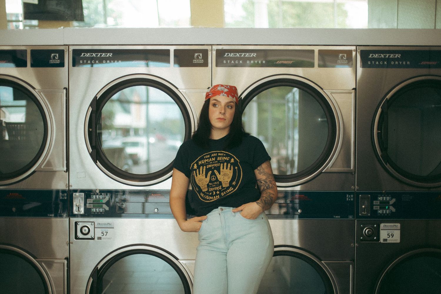 Two people pose casually in front of industrial washing machines at a laundromat.