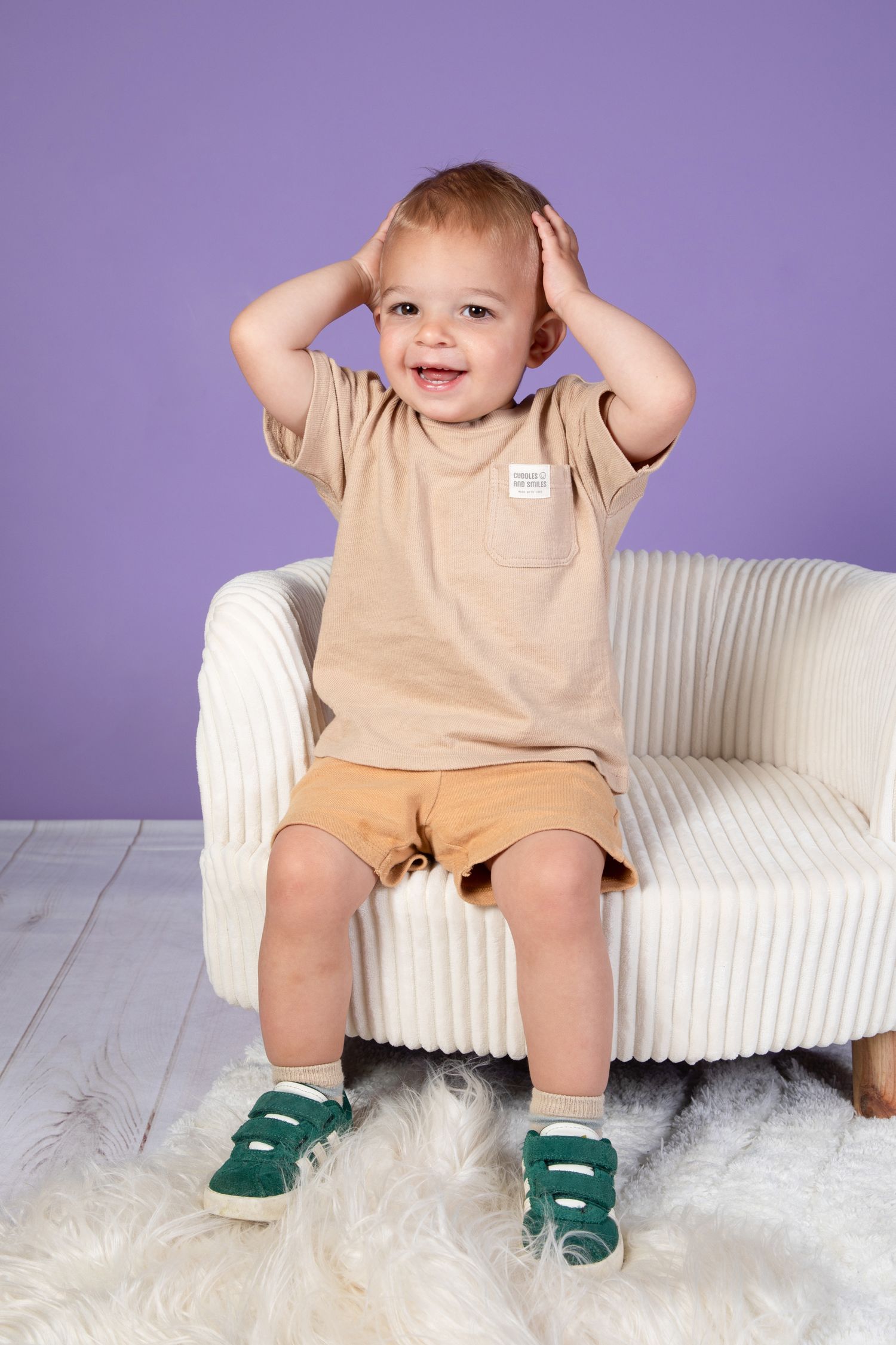 Small child in beige outfit and green socks sitting playfully on white modern chair.