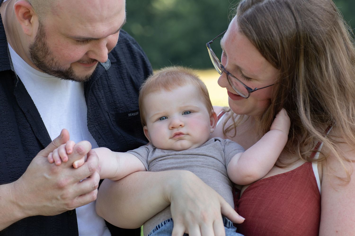 A bald father and blonde mother with glasses lovingly hold their blue-eyed baby outdoors in soft natural light.