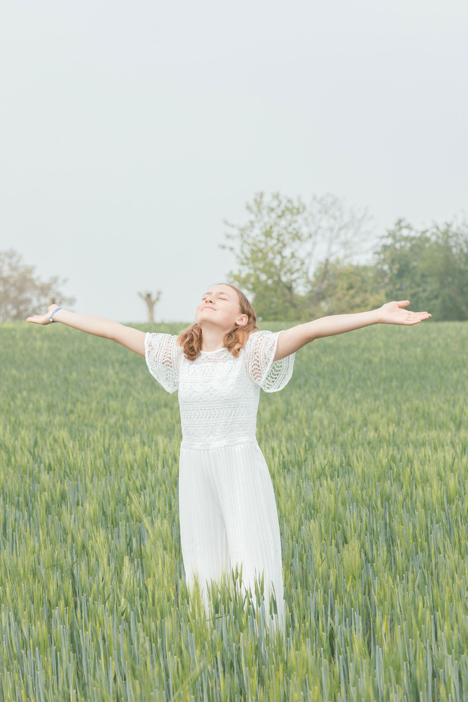 Figure in white dress stands joyfully with arms outstretched in green wheat field.