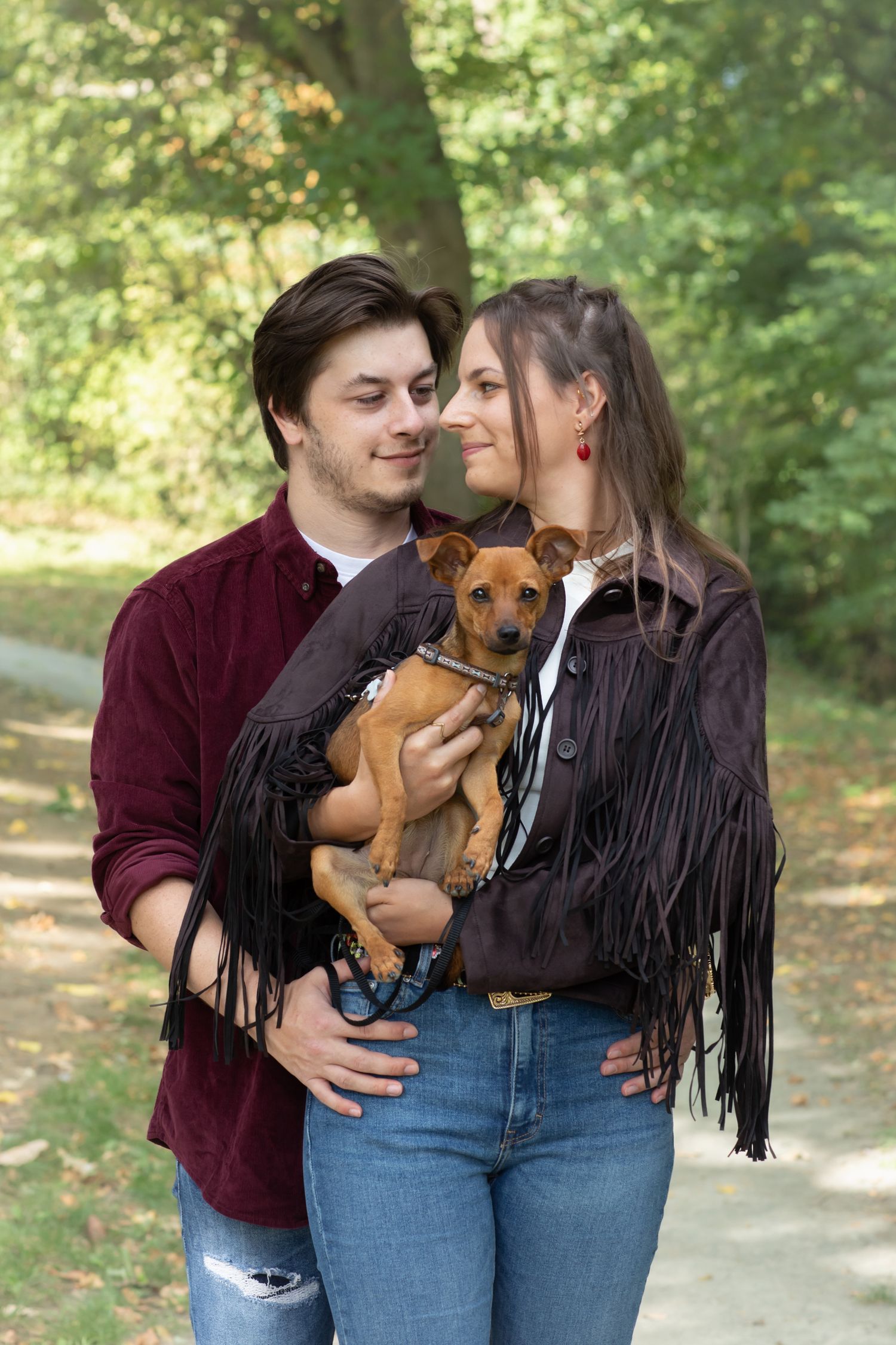 Couple in casual attire holding their brown dog while walking in a leafy park.