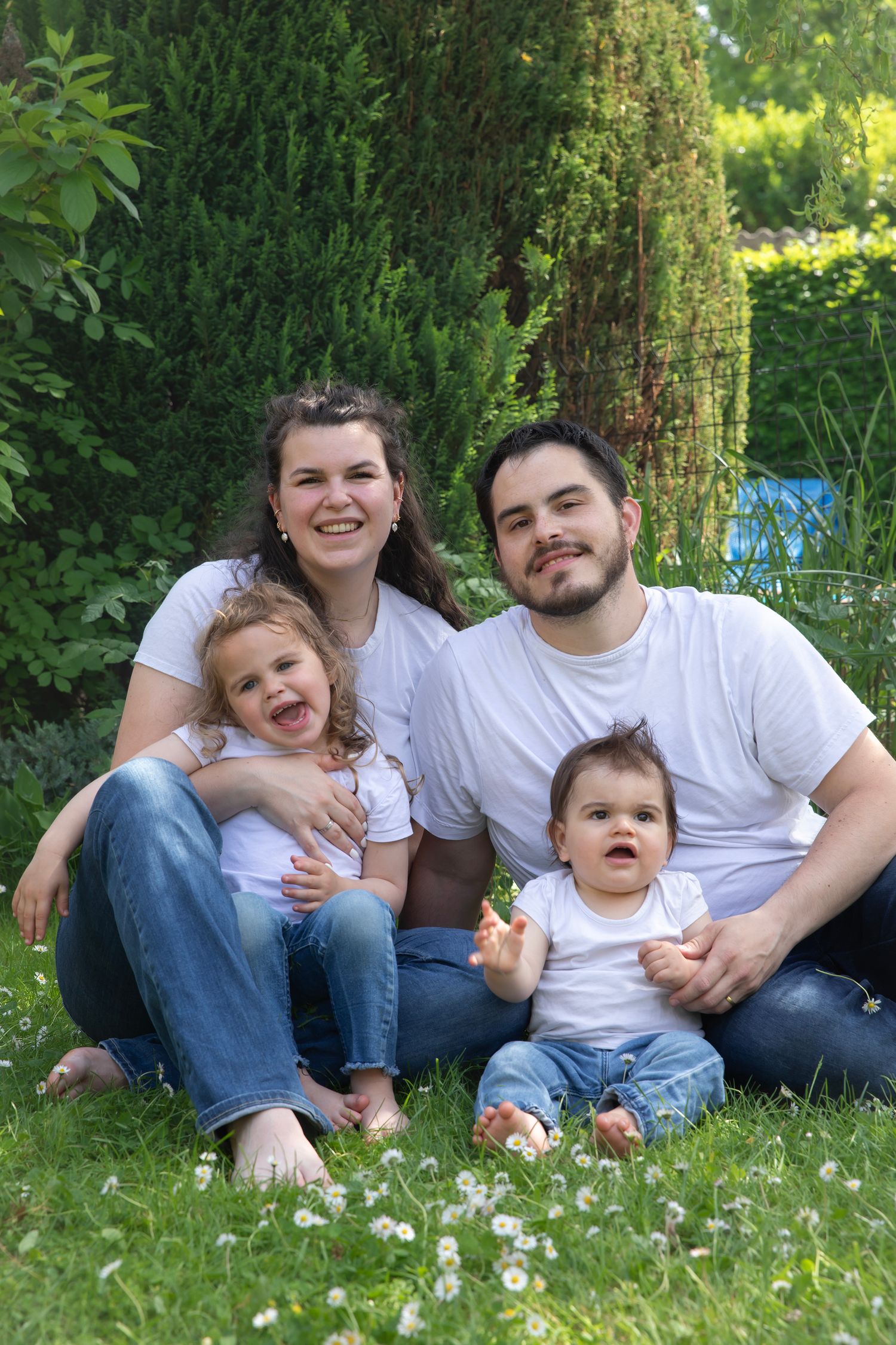 Happy family of four sitting on green grass, parents in white t-shirts and jeans with two young children outdoors.