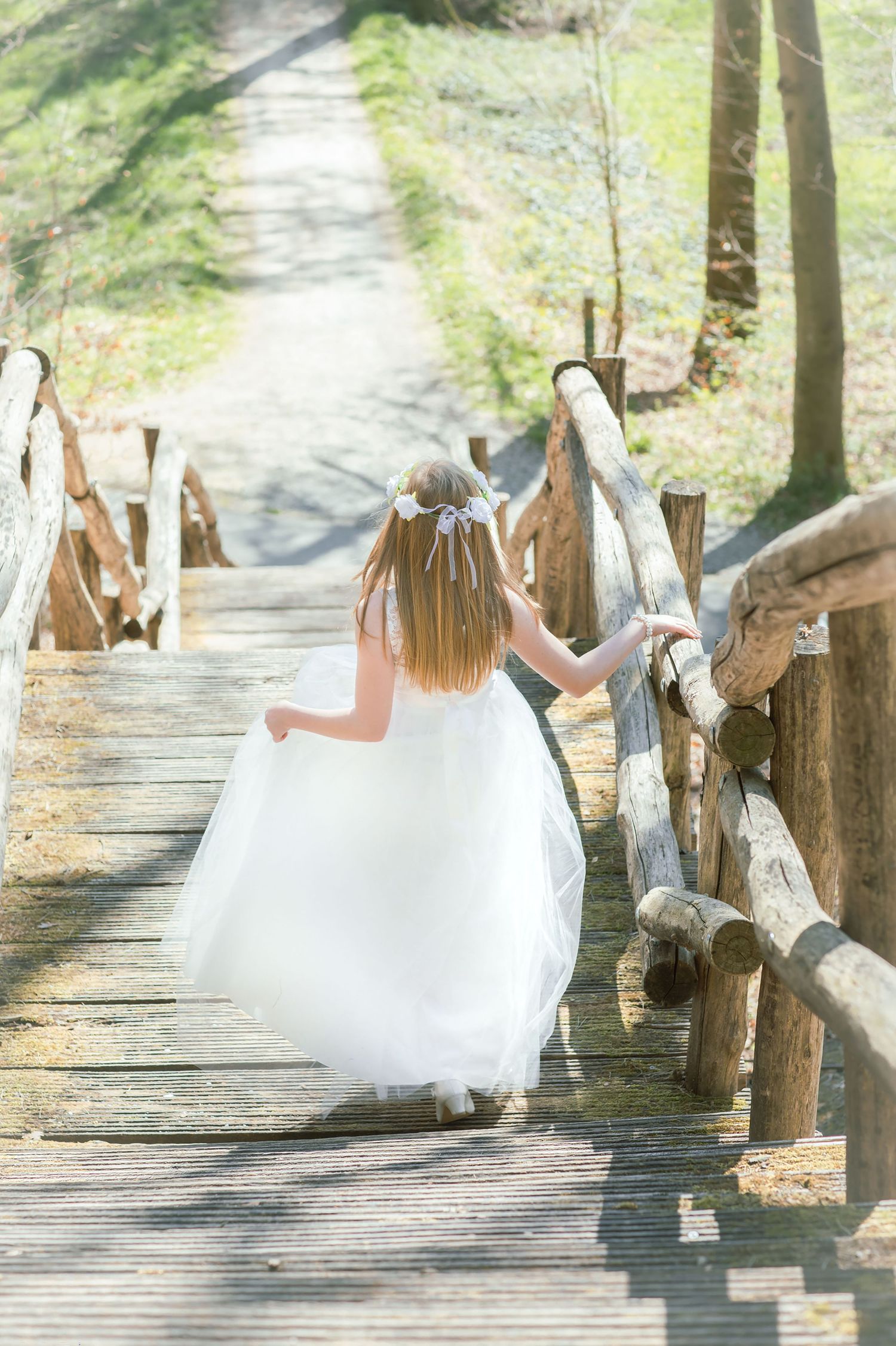 White dress flowing gracefully along a wooden boardwalk path through sunlit trees.