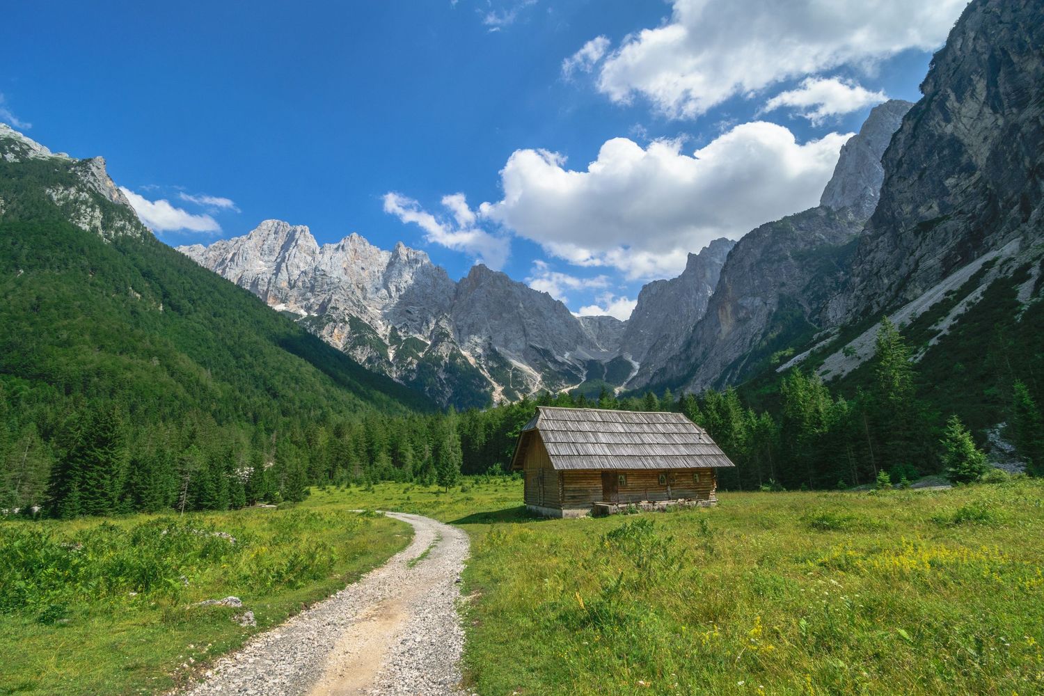 A rustic wooden cabin sits in an alpine meadow with a gravel path, surrounded by green forests and dramatic rocky mountains.