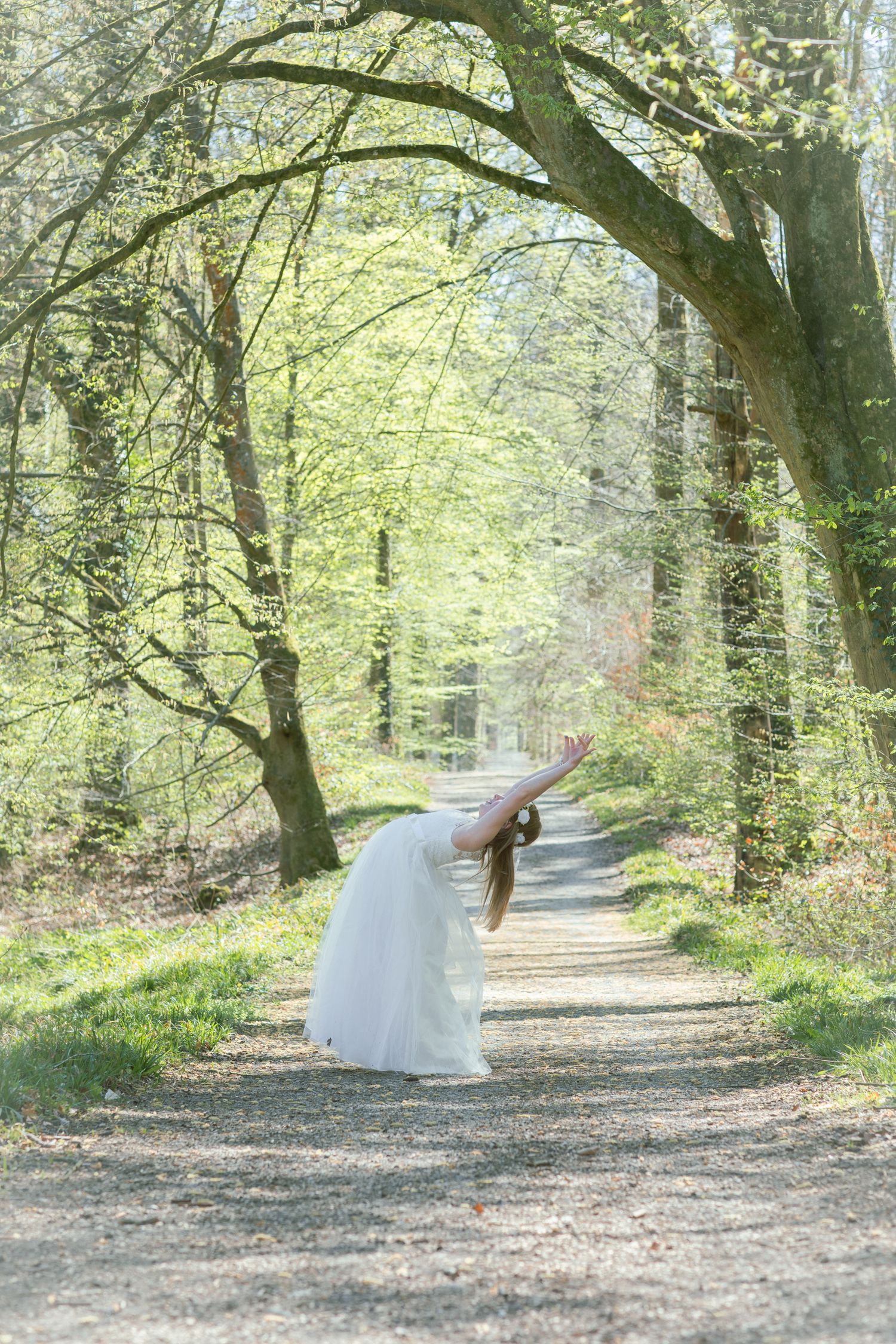 A graceful figure in a flowing white dress bends elegantly on a sunlit forest path surrounded by spring trees.