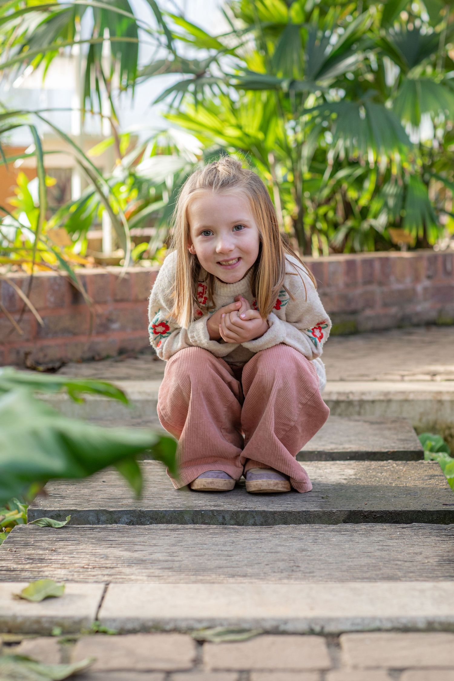 Child in pink outfit crouches playfully on brick pathway with greenery in background.