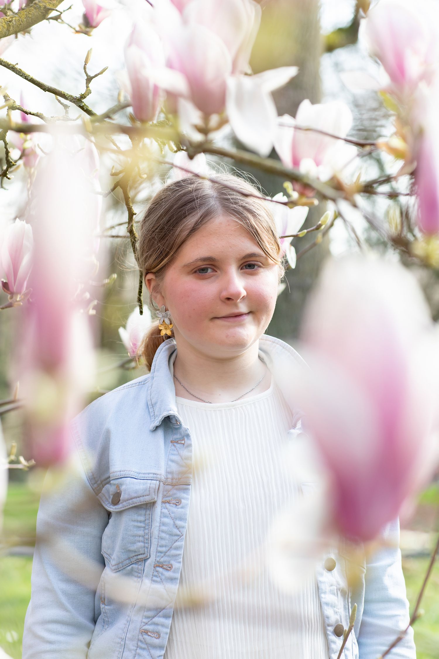 Young girl smiling among pink magnolia blossoms, wearing a denim jacket, in a sunlit spring garden.