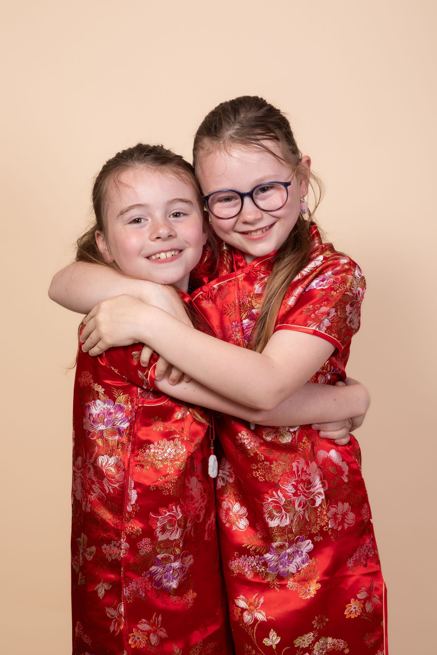 Two young girls in red floral Chinese qipao dresses hugging and smiling, one wearing glasses.