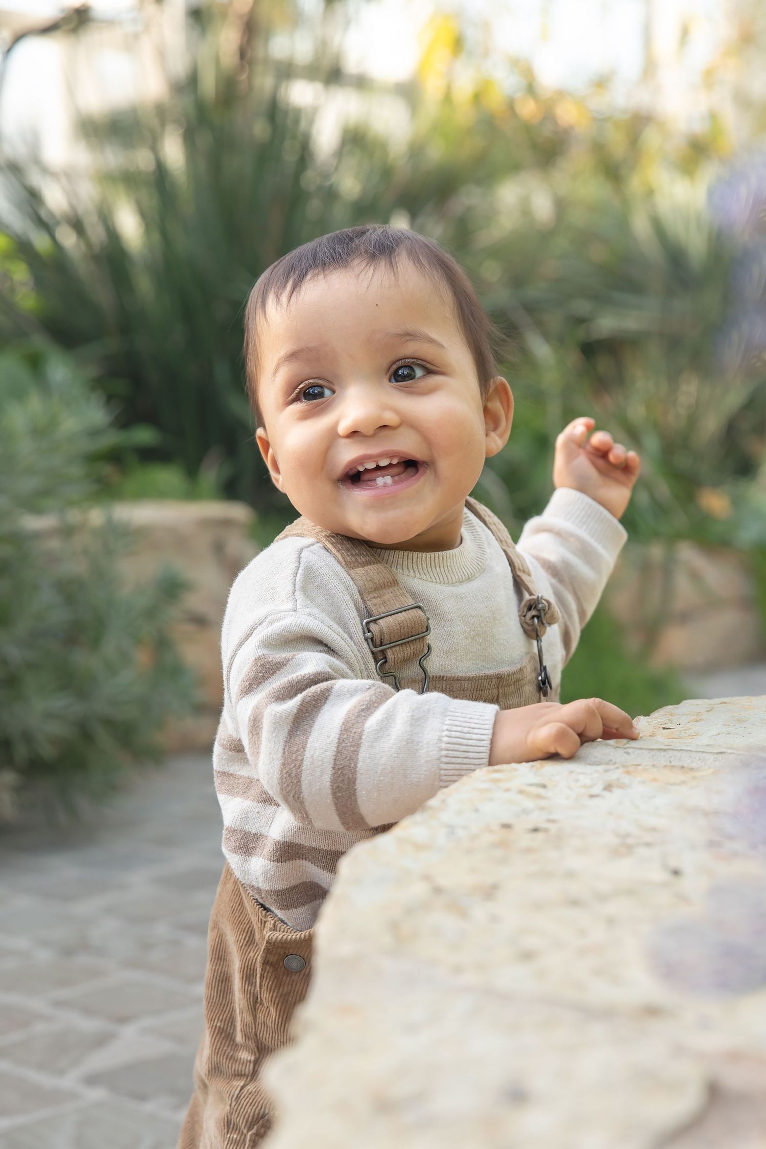Smiling baby boy in striped sweater and overalls standing at a stone ledge outdoors in a garden.