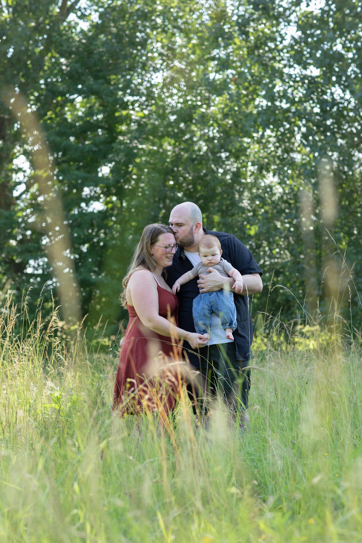 A couple stands in a lush green meadow, the man kissing the woman's head while holding their baby.