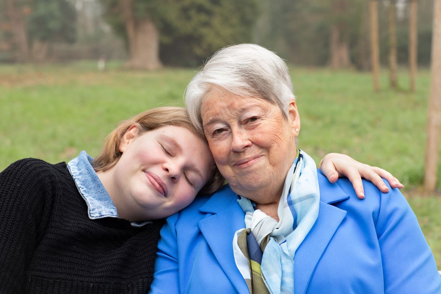 A young girl rests her head on an elderly woman's shoulder outdoors, both smiling warmly in a park setting.