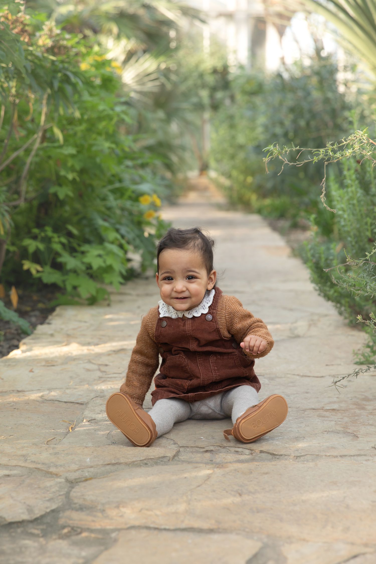 A baby in a brown sweater and white pants sits on a stone pathway surrounded by greenery.