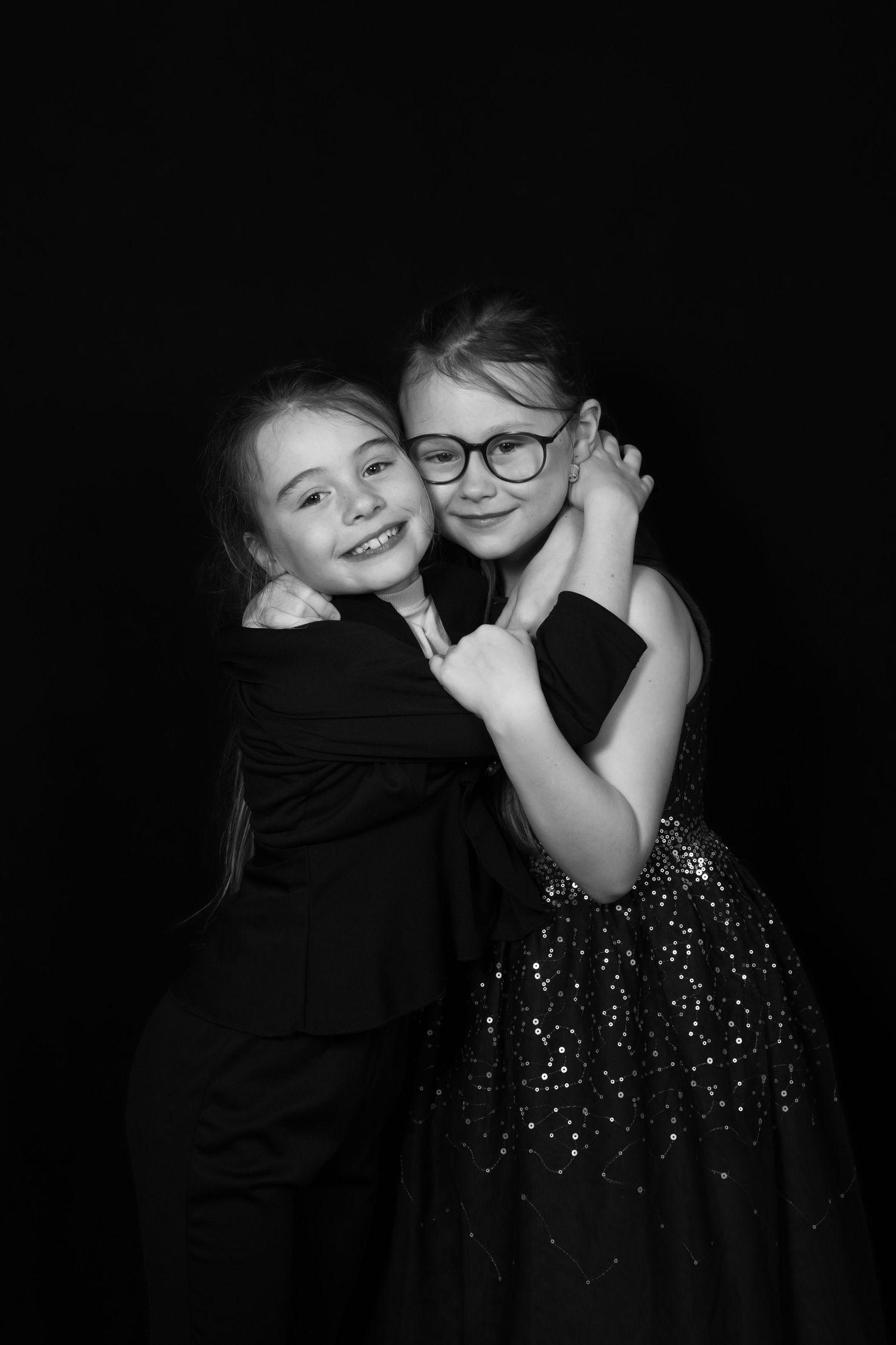Two young girls in elegant black outfits hugging and smiling in a black and white studio portrait photo.