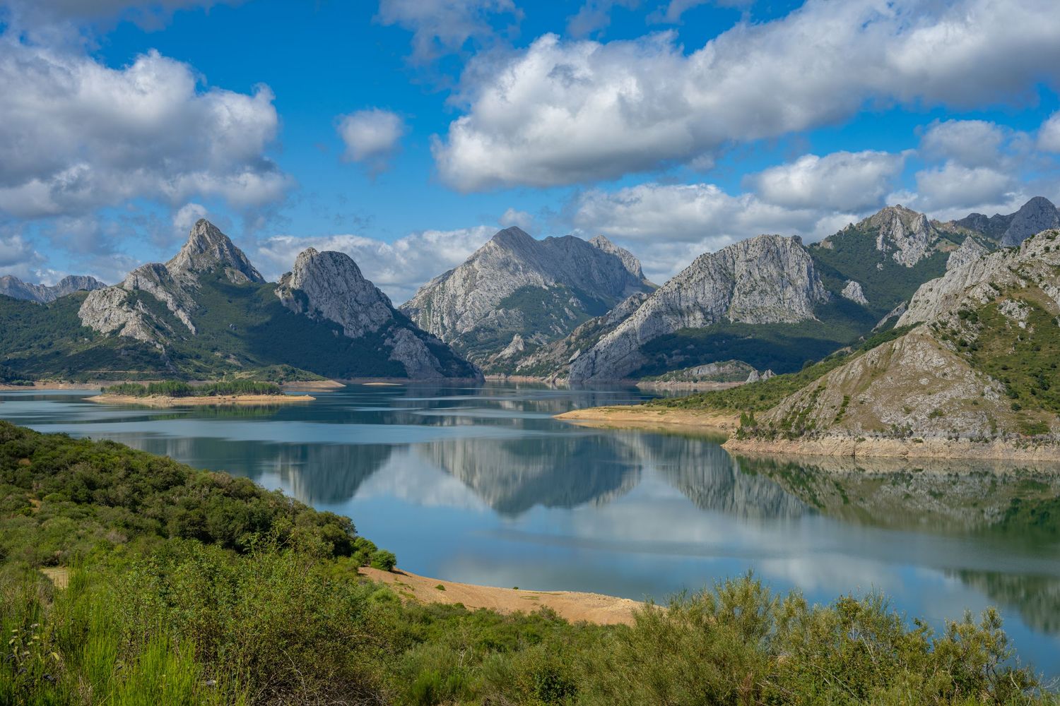 Scenic mountain lake reflecting peaks under blue sky with white clouds, surrounded by lush green vegetation in Spain.