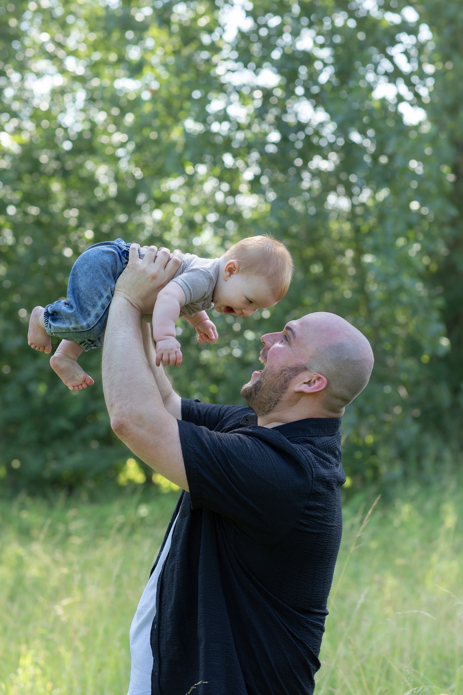 Bald father lifting happy baby in the air outdoors in a green park, sharing a joyful moment together.