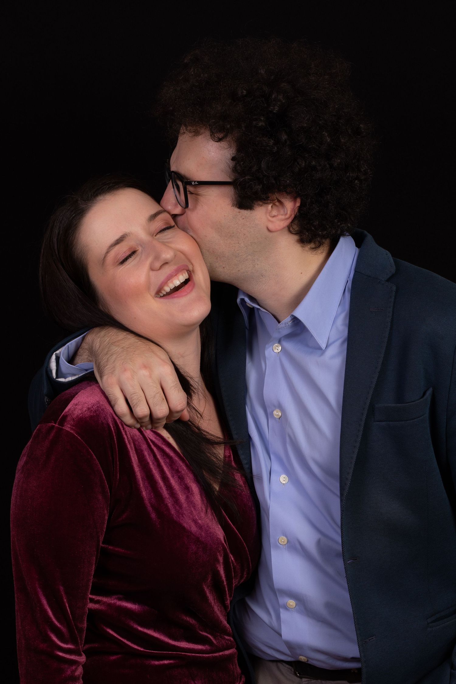 A couple shares a tender moment against a dark background, with one wearing a burgundy velvet top and the other in a navy blazer.