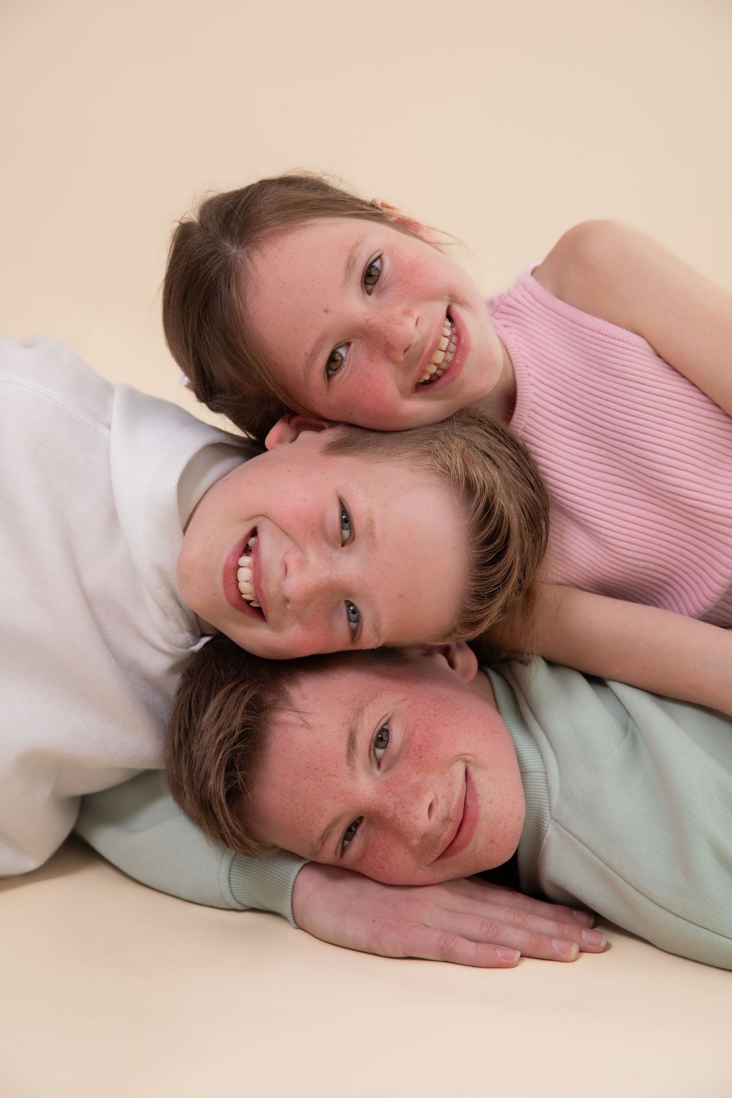 Three smiling children stacked together, lying down and posing for a fun portrait photo against a beige background.