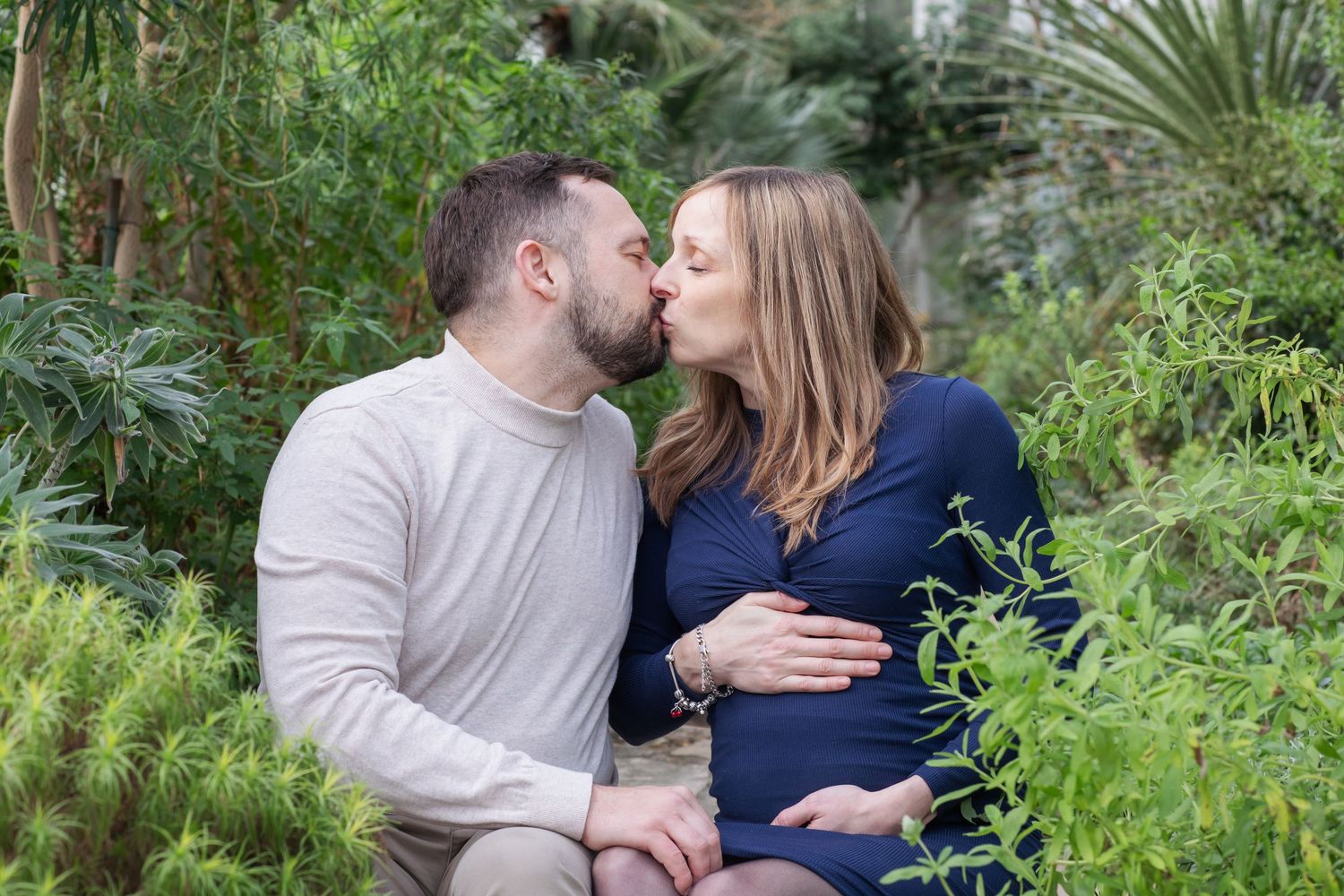 Expectant couple sharing tender moment in lush greenhouse setting.