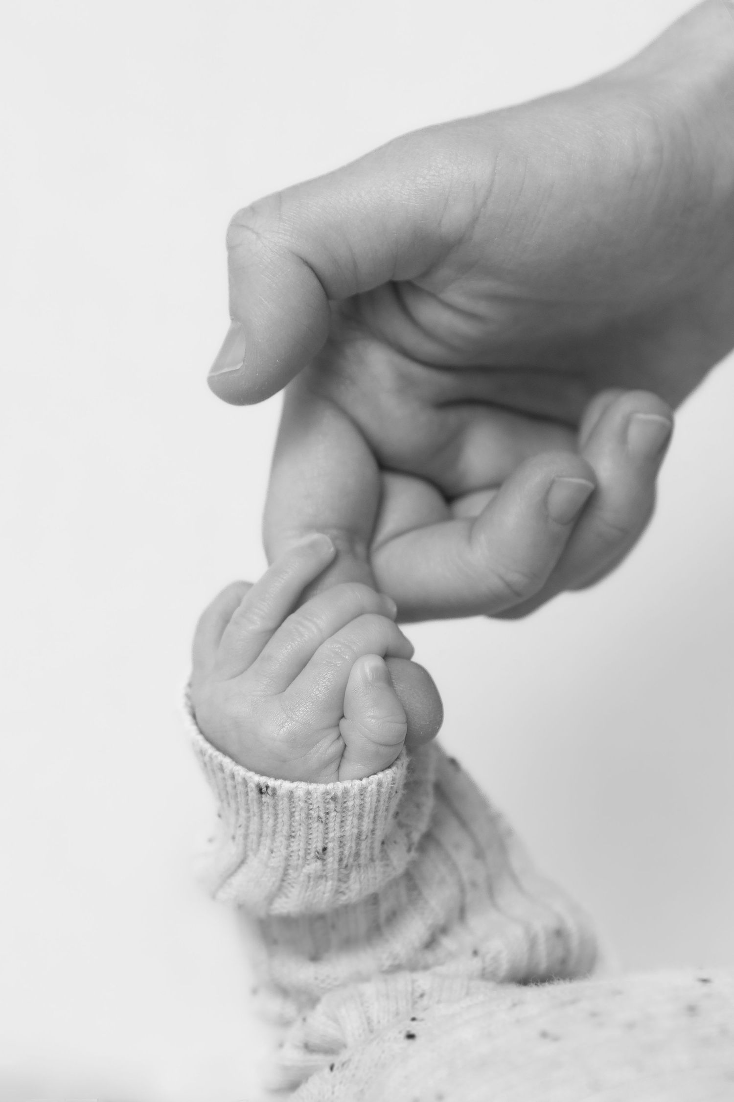 Black and white photo of a newborn baby hand grasping an adult finger, wearing a knit sleeve.