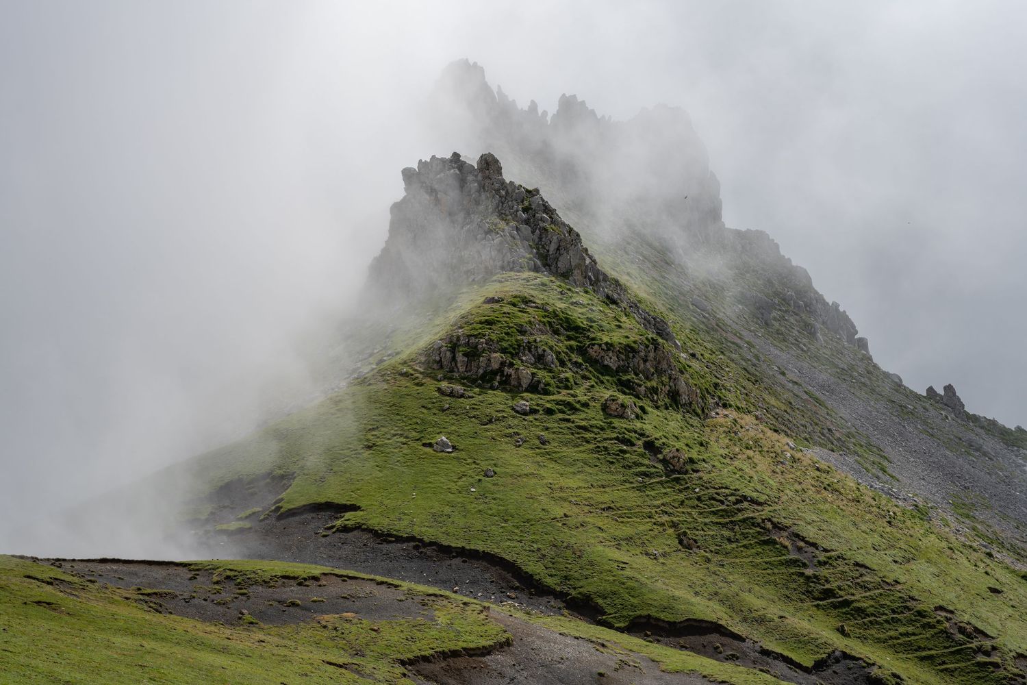 Misty green mountain peak shrouded in clouds with rocky summit and lush moss-covered slopes.