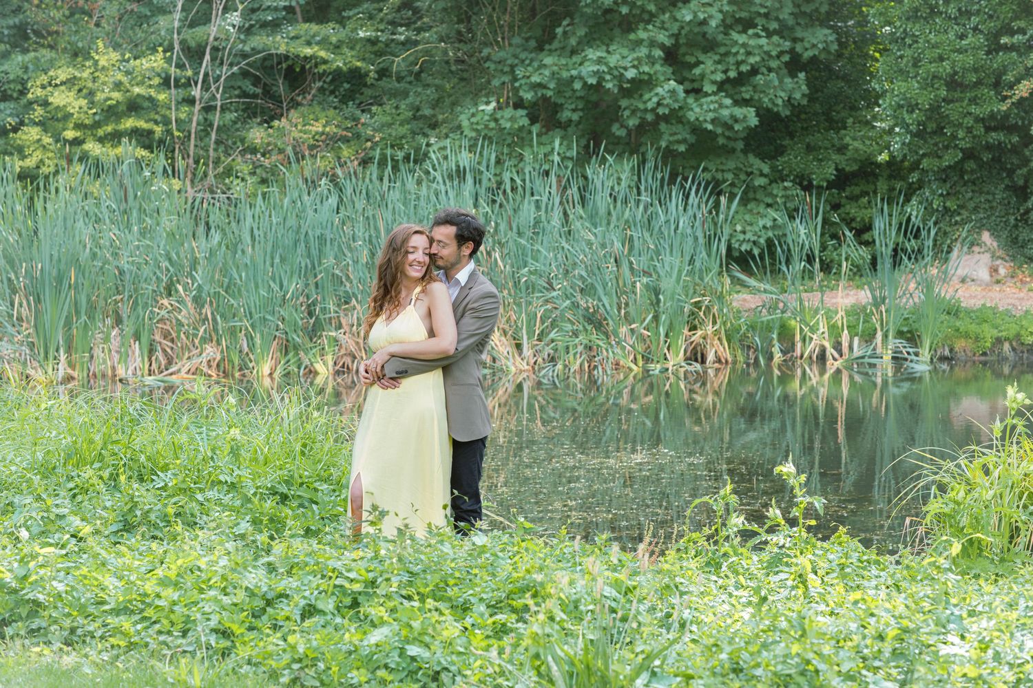 Couple embracing romantically by a serene pond surrounded by lush green foliage and tall reeds.