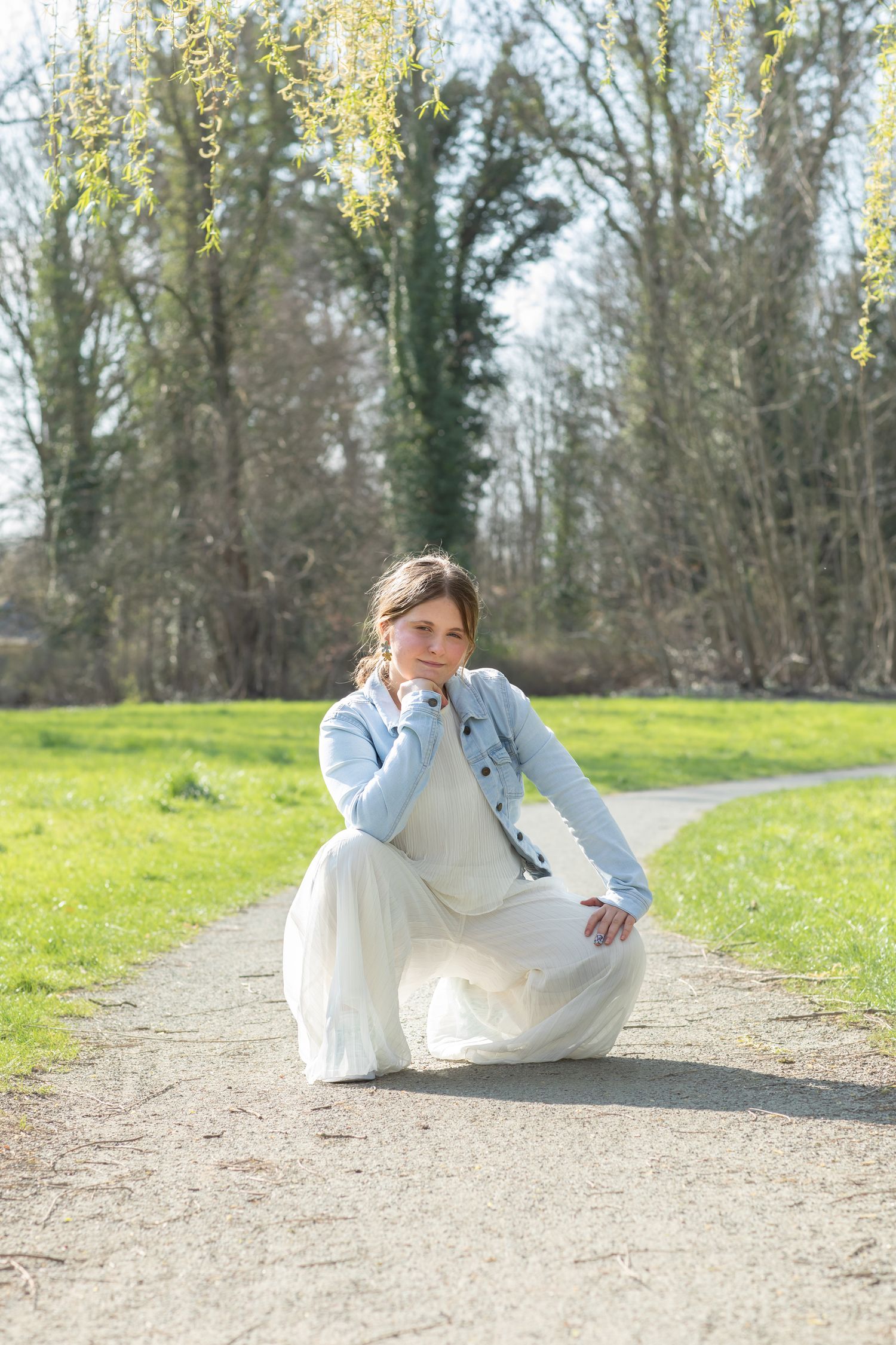 Young woman in white dress and denim jacket crouching on a park path, surrounded by green trees on a sunny day.