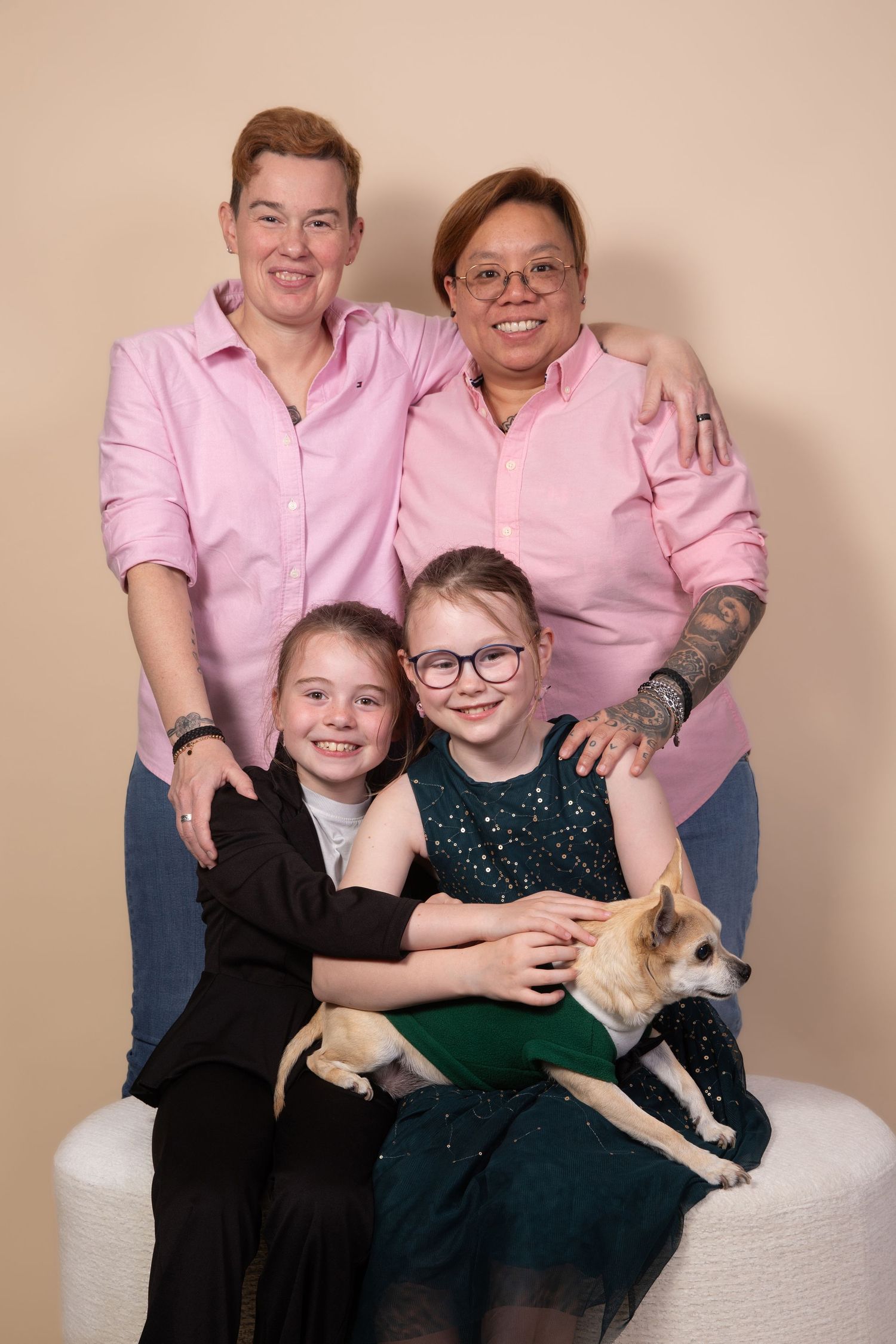 Family portrait of two women in pink shirts with two girls and a small tan dog against a beige background.