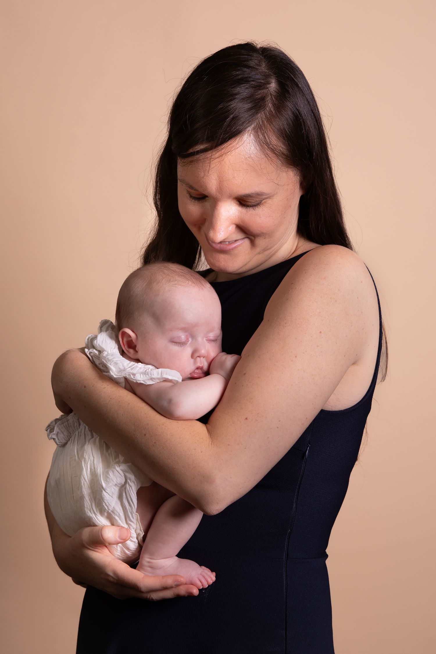 A tender moment between parent and sleeping newborn baby dressed in white against a neutral beige background.