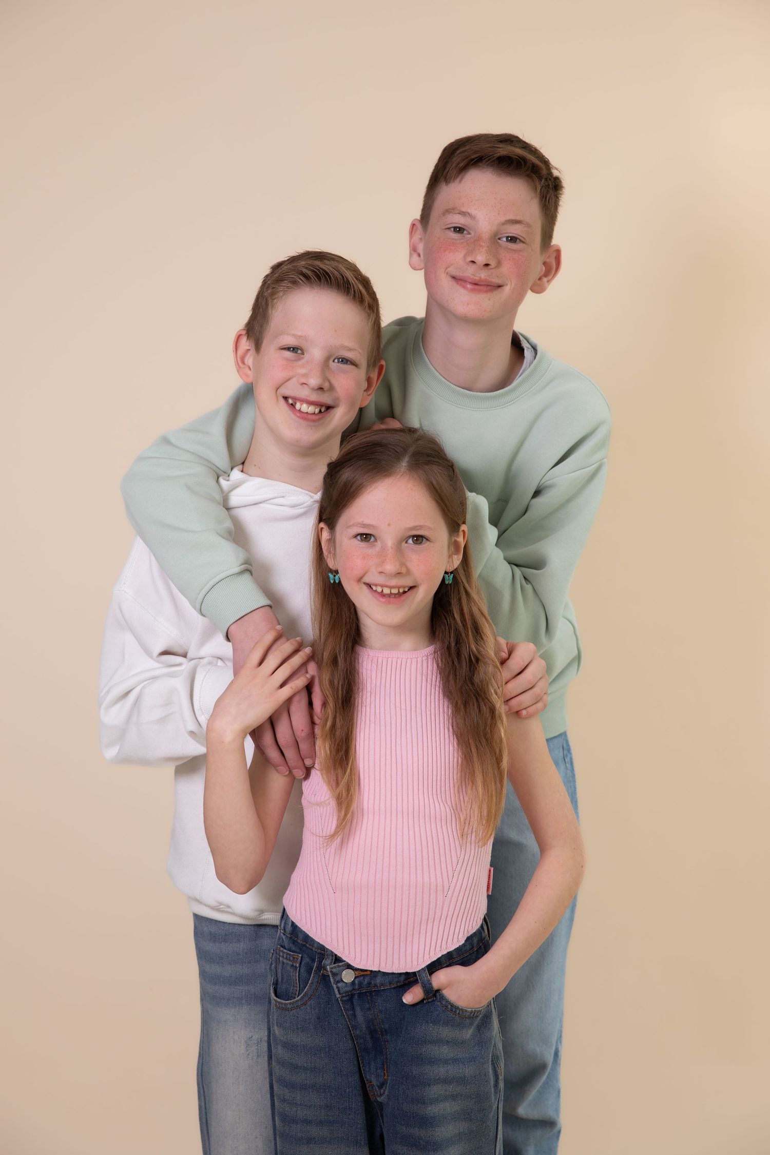 Three smiling siblings pose together against a beige background, wearing casual jeans and colorful tops.