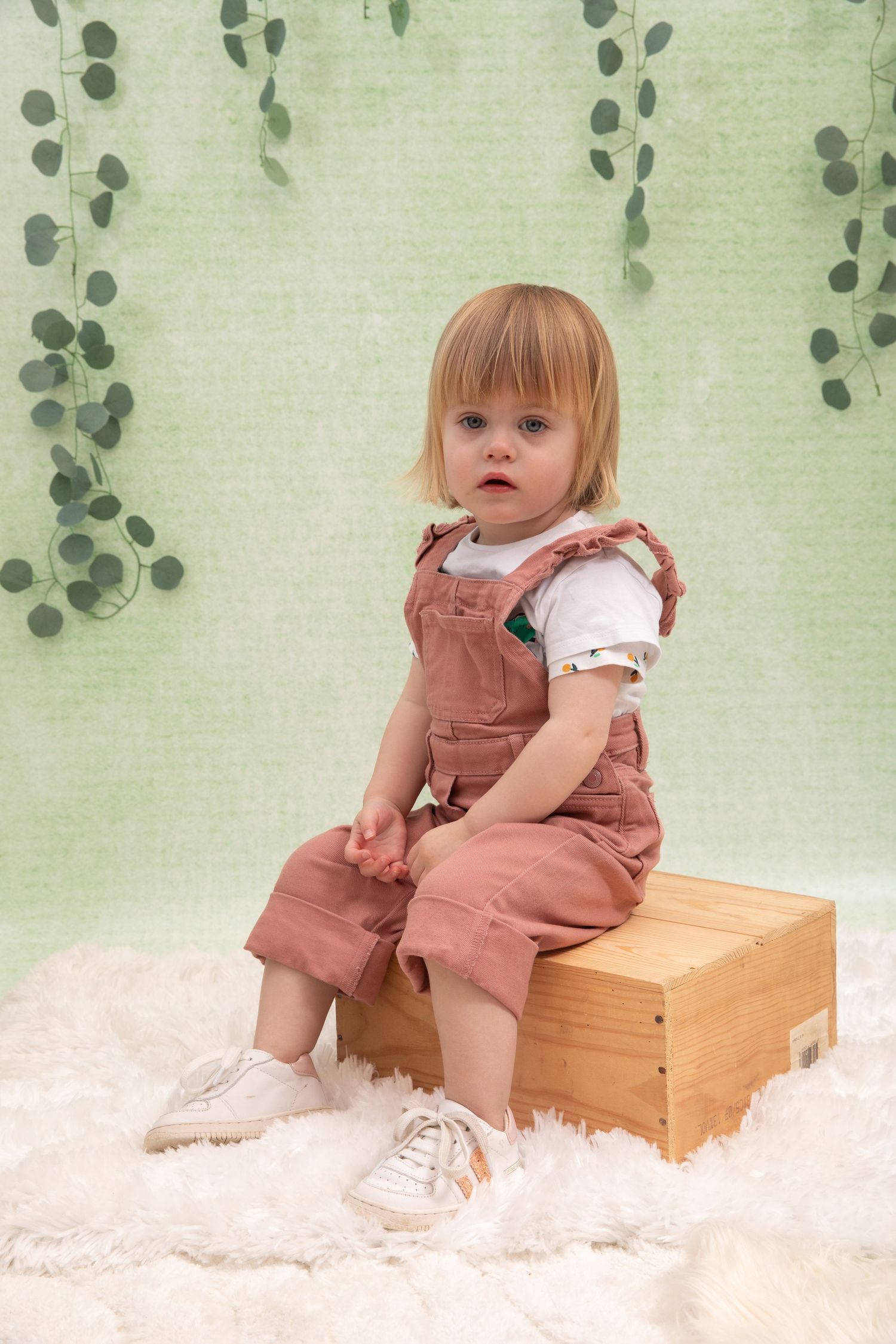 Toddler girl in pink overalls sitting on wooden box against green backdrop with eucalyptus garlands.