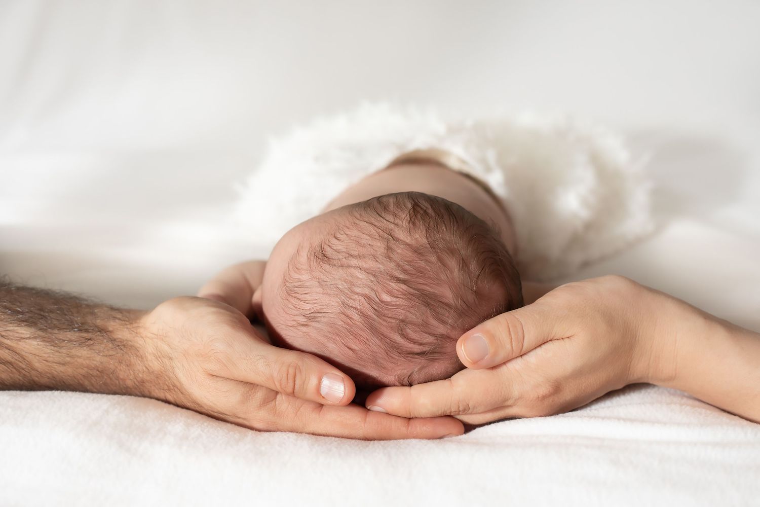 Adult hands gently cradling a newborn baby on white bedding.