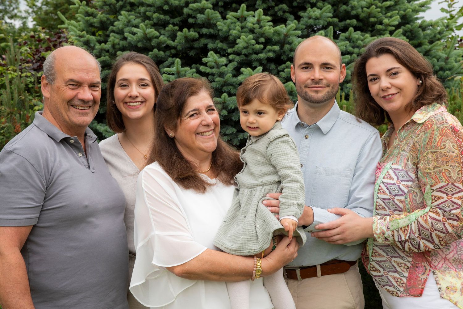 Multi-generational family of six smiling together outdoors, including grandparents, parents, and a toddler girl.