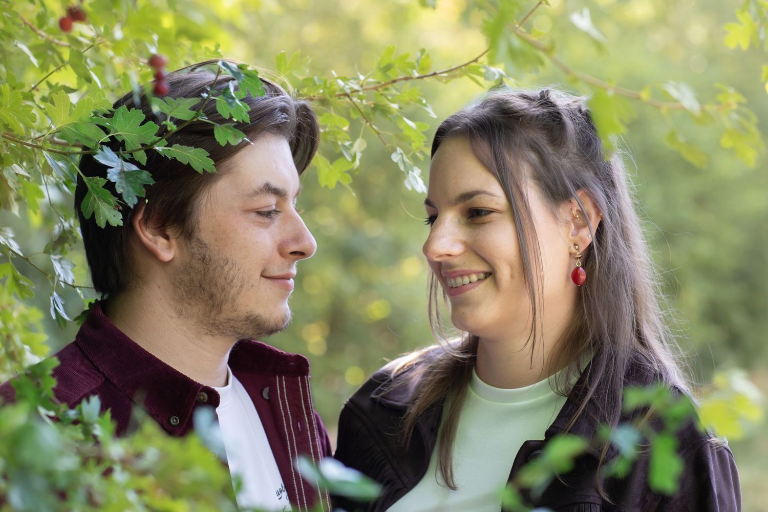 A couple shares a tender moment outdoors among green foliage, with one wearing a leaf crown in their hair.