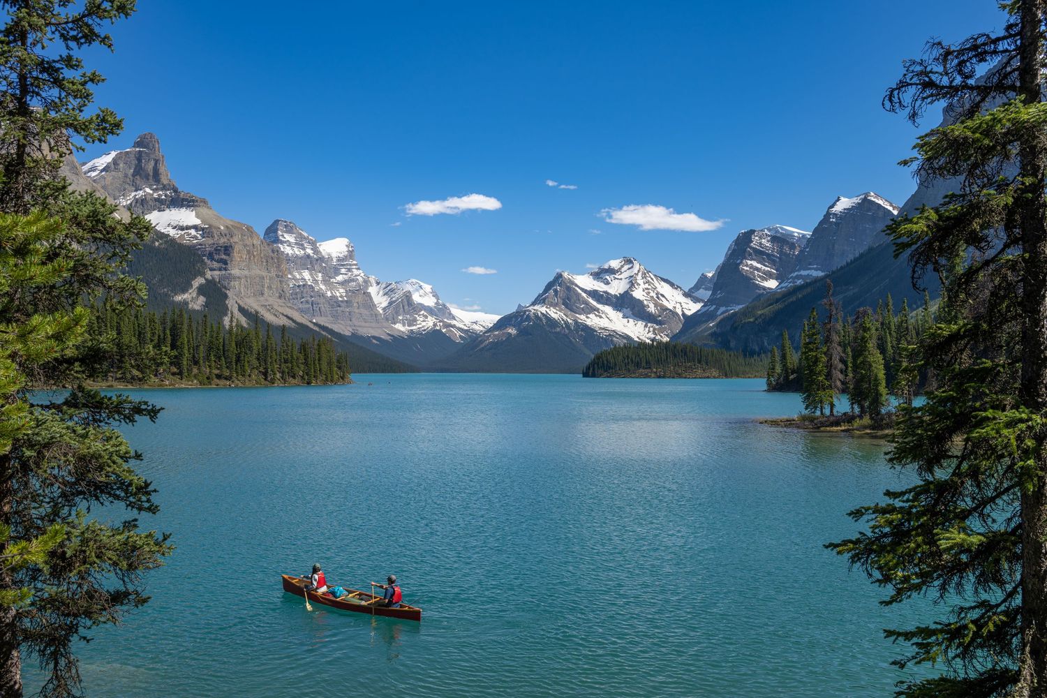 Two people canoe on turquoise Maligne Lake surrounded by snow-capped Canadian Rocky Mountains under a clear blue sky.