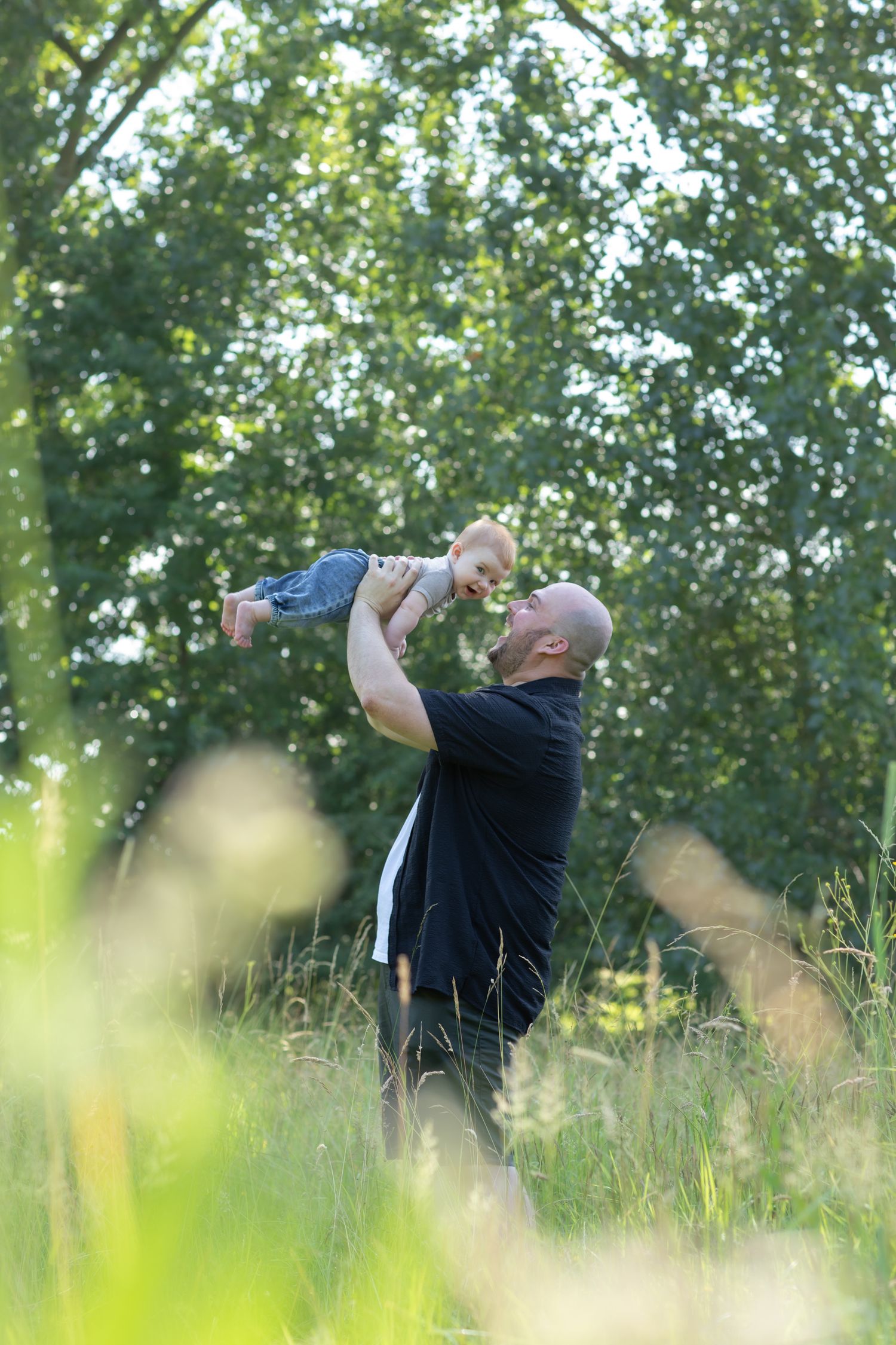 A bald father lifts his smiling baby overhead in a sunny green meadow with tall grass and trees behind him.